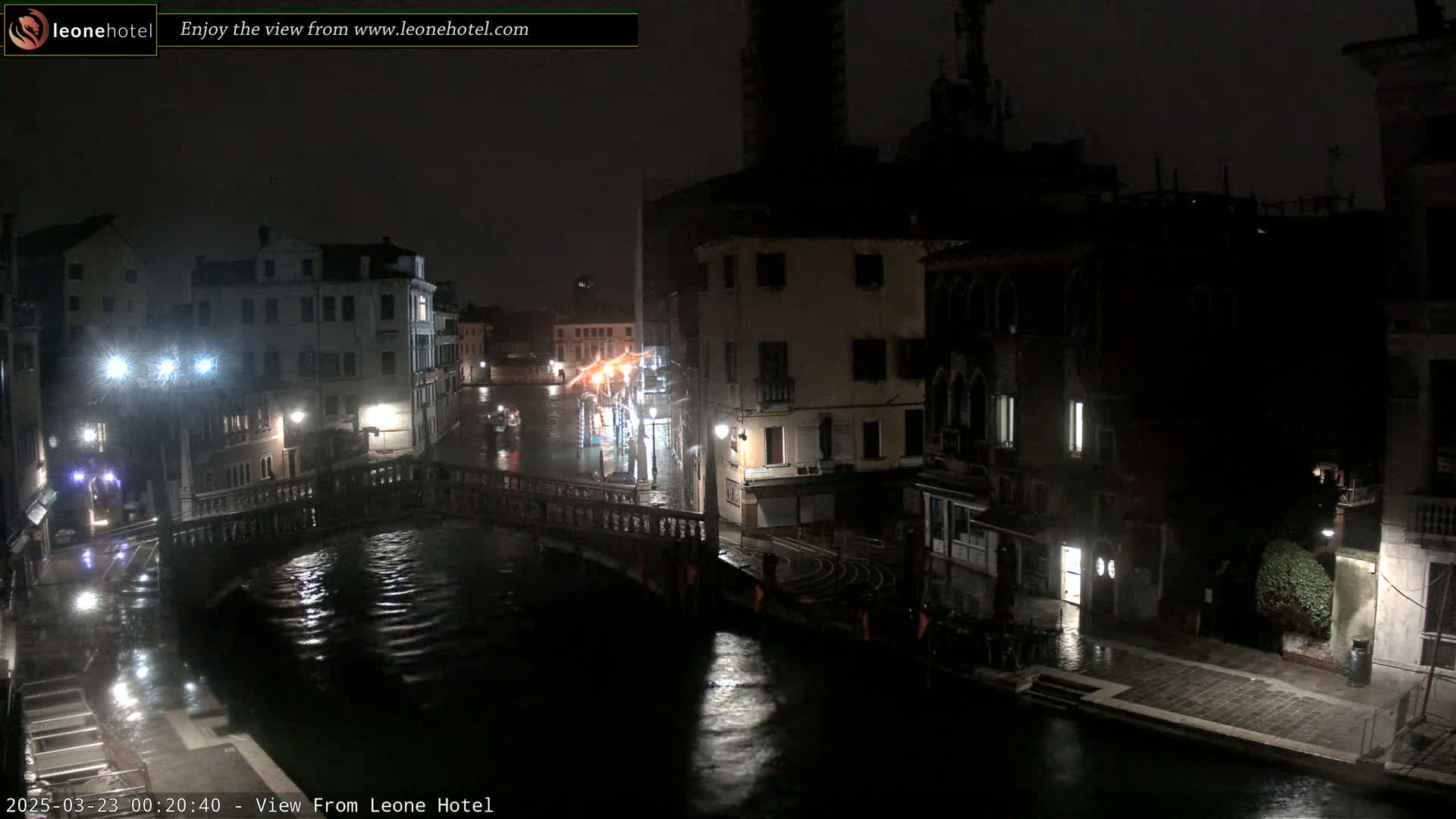 A nighttime view of a rain-slicked canal in Venice, showing several buildings along the waterway and a bridge crossing the canal.