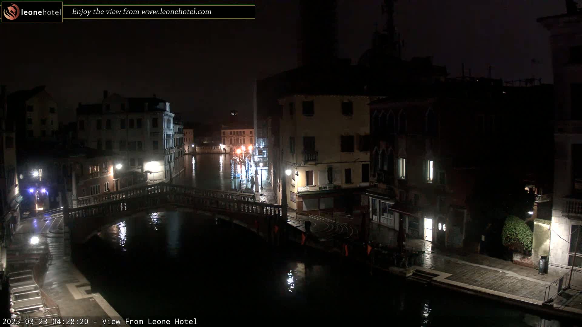 A nighttime view of a canal in Venice, Italy, shows a bridge and buildings illuminated by streetlights under a dark sky with wet surfaces.