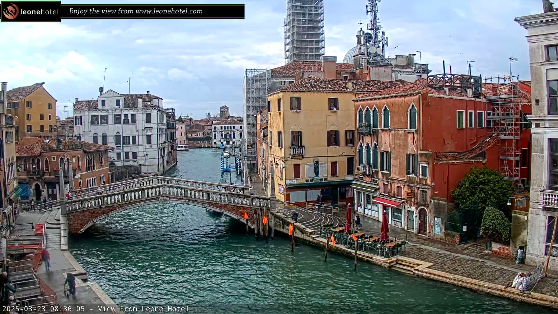 A canal in Venice, Italy, with several colorful buildings lining its banks, a stone bridge spanning the waterway, and a cloudy sky overhead.