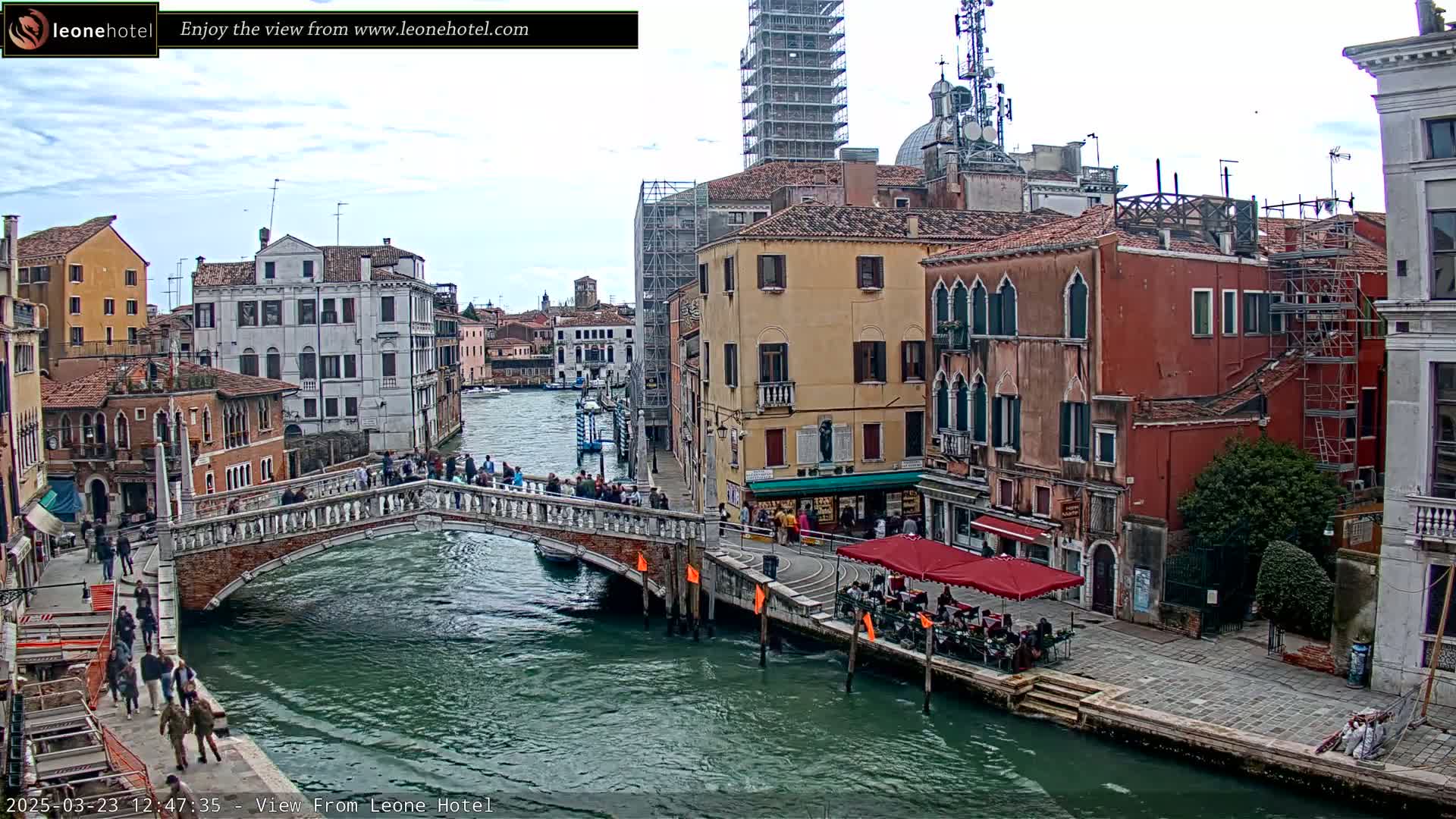 A canal in Venice, Italy, under a partly cloudy sky, is spanned by a bridge with people walking across it and lined with historic buildings, one of which is under construction.