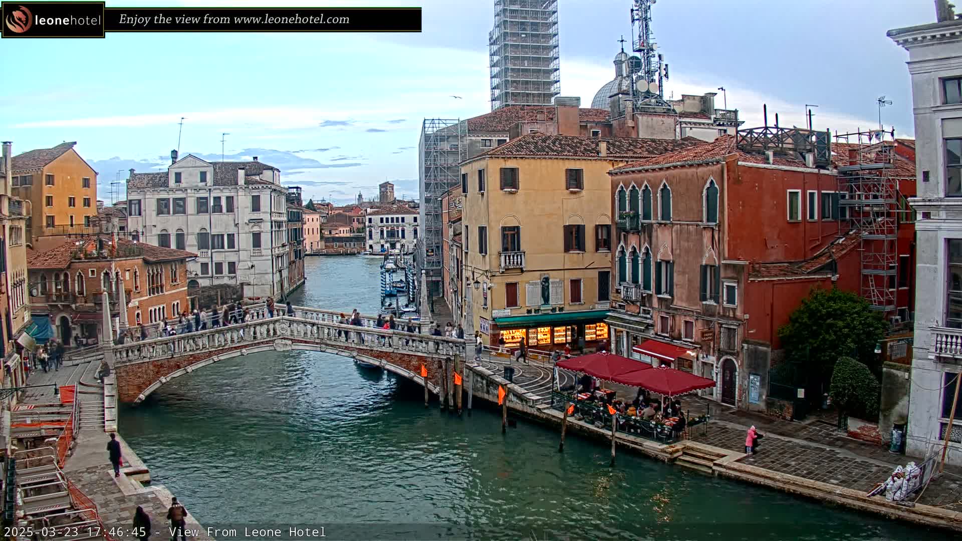 A stone bridge crosses a canal in Venice, Italy, under a partly cloudy sky, with pedestrians walking across the bridge and people sitting at an outdoor cafe along the canal.
