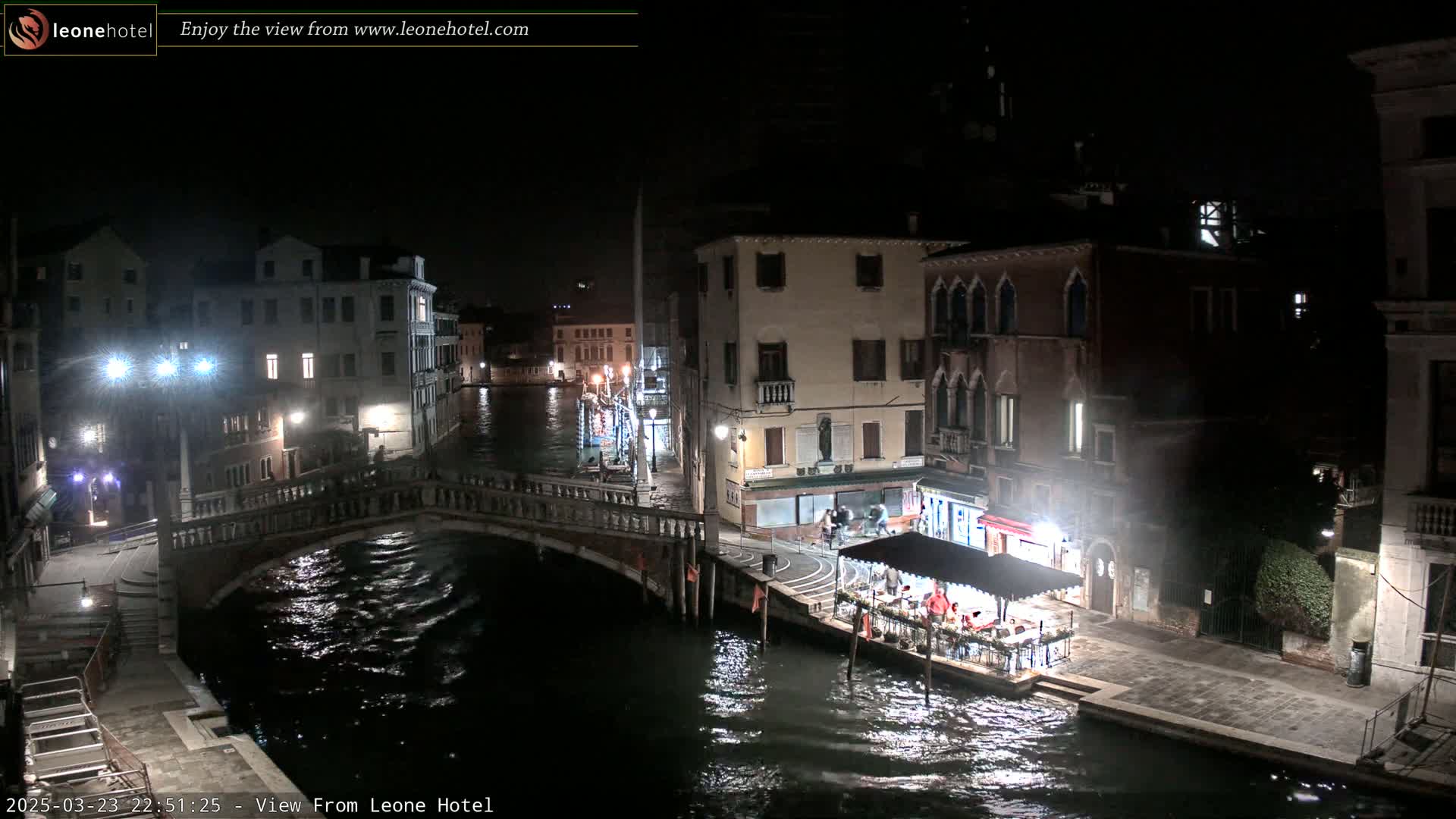 A nighttime view of a canal in Venice, with bridges, buildings, and a small restaurant visible along the waterway.