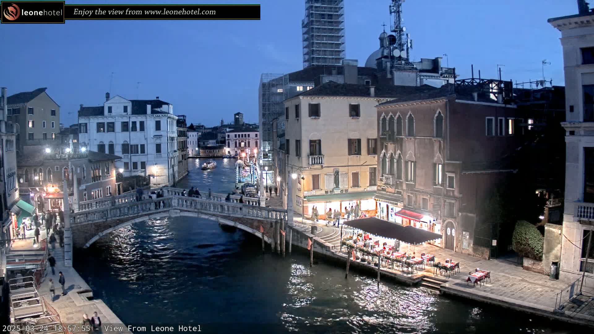 A twilight view of a canal in Venice, showing a stone bridge with pedestrians, buildings lining the waterway, and an outdoor restaurant with diners.