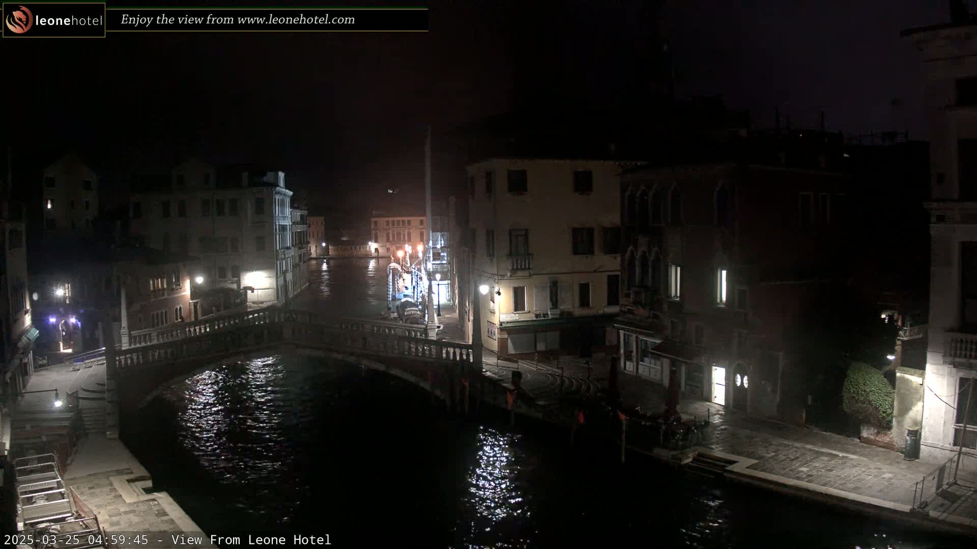 A nighttime view of a canal in Venice, Italy, shows a bridge and buildings lining the waterway under a dark sky.