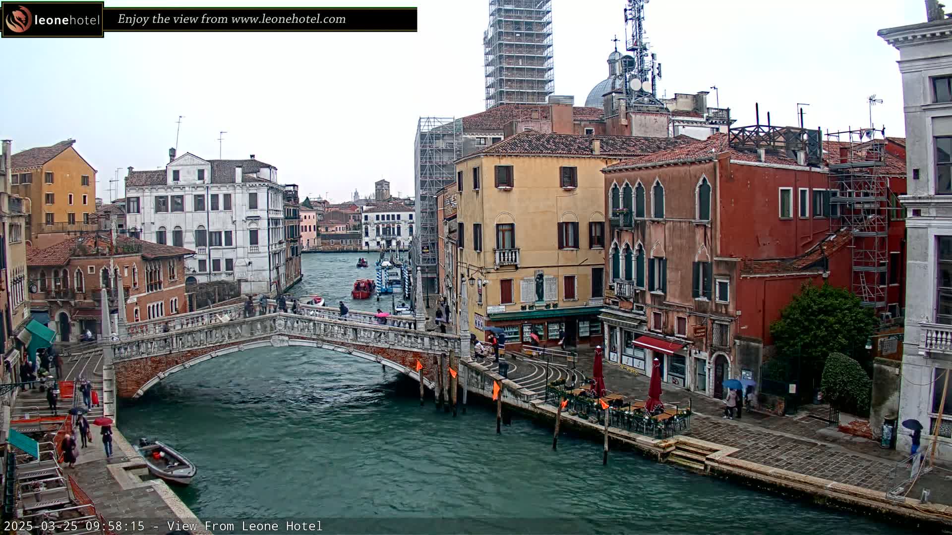 A stone bridge arches over a canal in Venice, Italy, under an overcast sky, with colorful buildings lining the waterway and several people walking along the canal's edge.