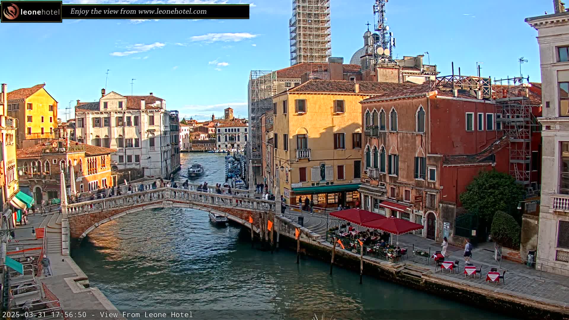 A sunny day in Venice shows a canal with a stone bridge crowded with pedestrians, colorful buildings lining the waterway, and several boats passing by.
