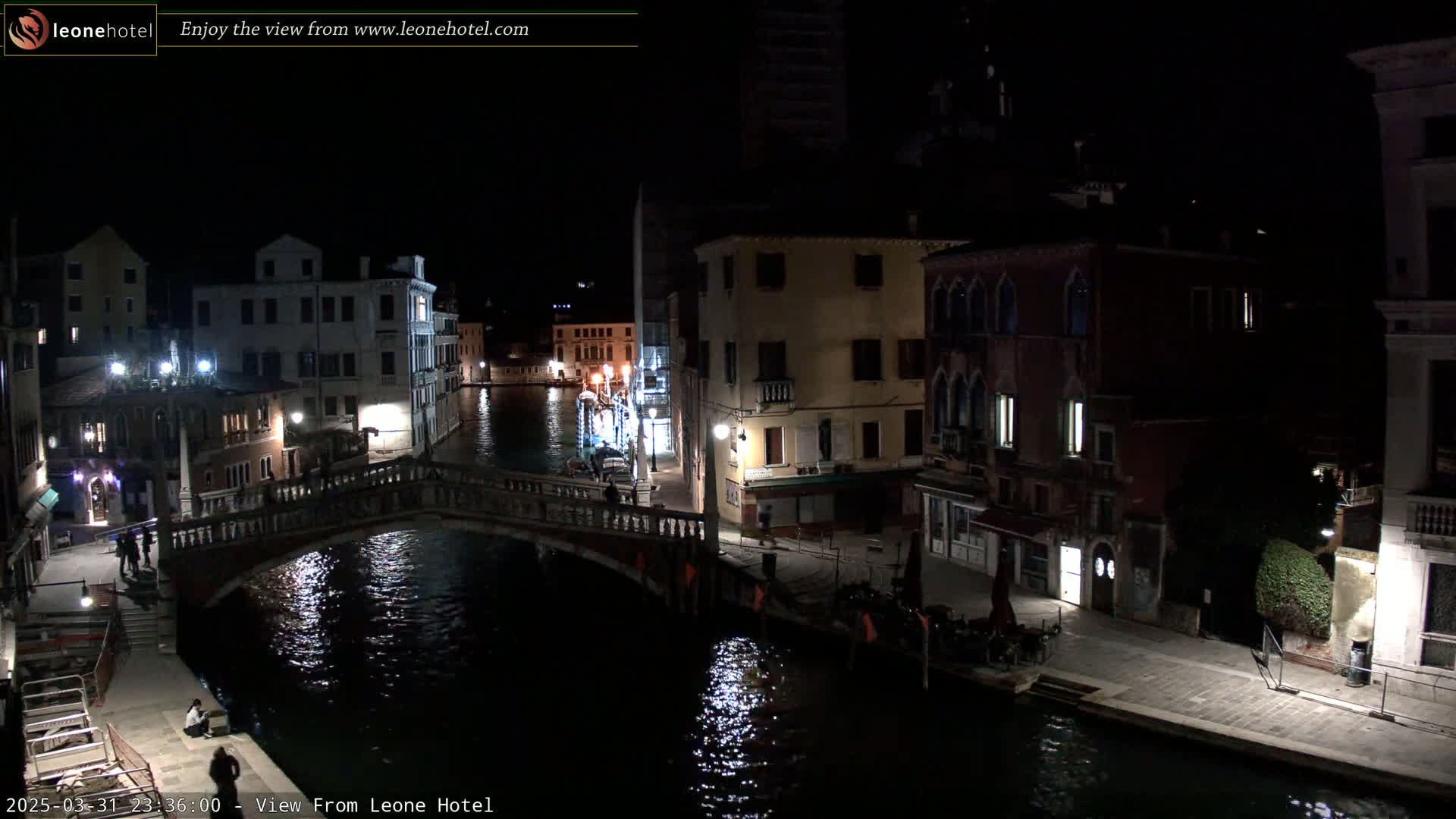 A nighttime view of a canal in Venice, Italy, with bridges, buildings, and a few people visible along the waterway.