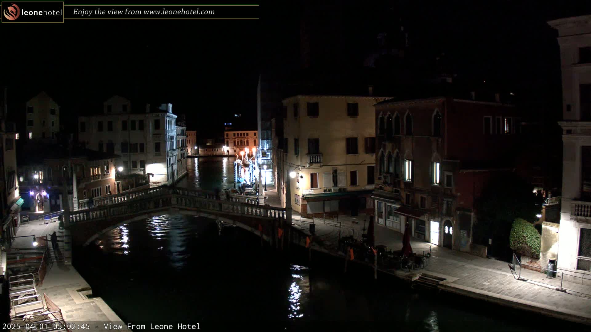 A nighttime view of a canal in Venice, Italy, shows a bridge over the water with buildings lining the waterway, some with lights on.