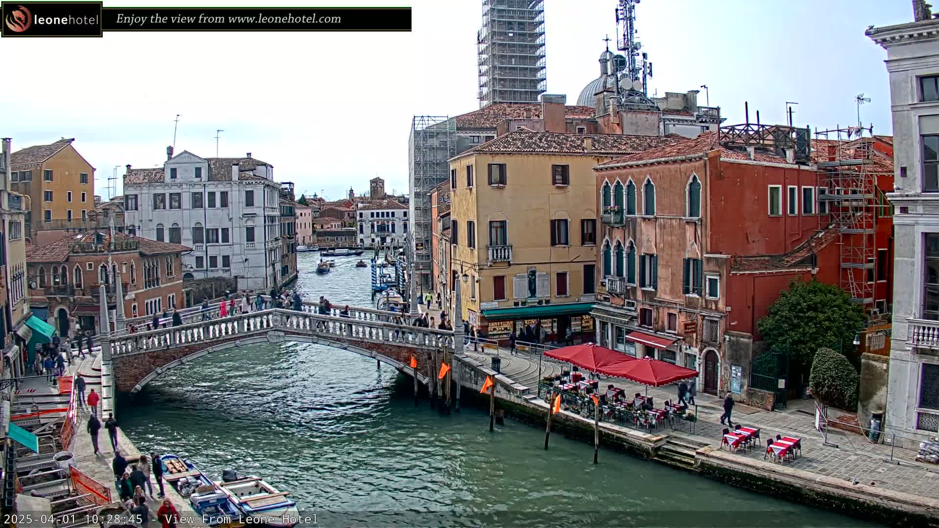 A canal in Venice, Italy, under a mostly cloudy sky, is spanned by a bridge with pedestrians, and lined with buildings, some under construction, and outdoor seating at a restaurant.