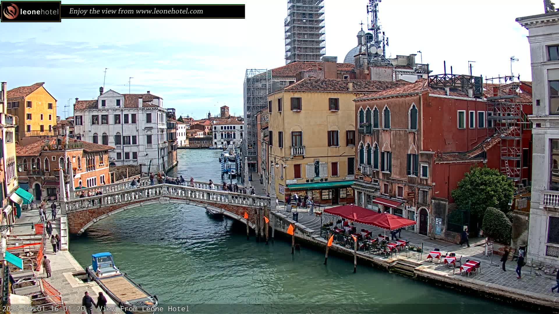 A canal in Venice, under a partly cloudy sky, is spanned by a stone bridge with pedestrians crossing it, and lined with colorful buildings, one of which has outdoor seating.