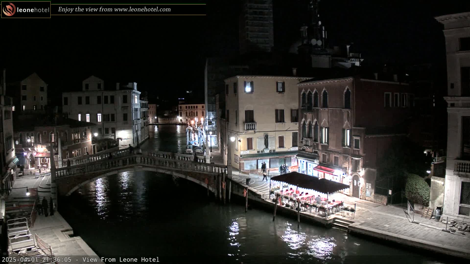 A nighttime view of a canal in Venice, Italy, shows a bridge, buildings lining the waterway, and an outdoor restaurant with patrons under a canopy.