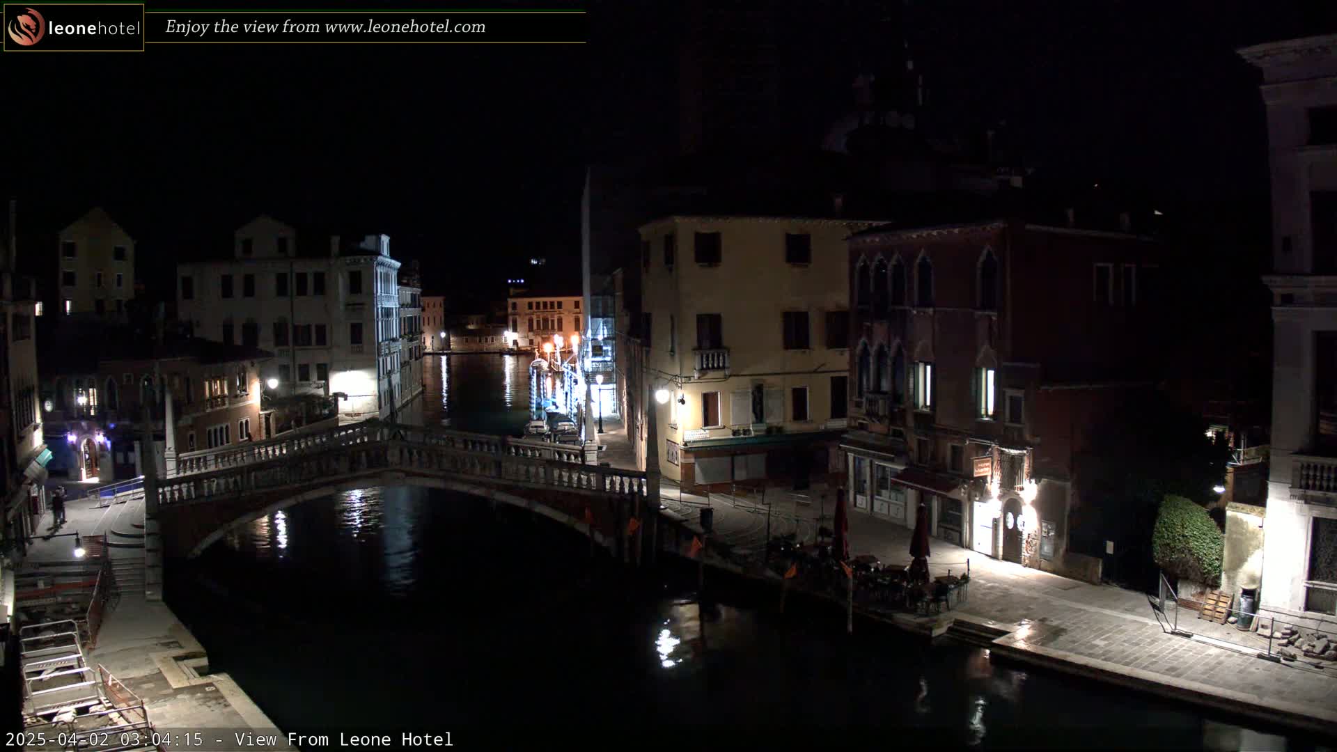 A nighttime view of a canal in Venice, Italy, shows a stone bridge, buildings lining the waterway, and some outdoor seating.