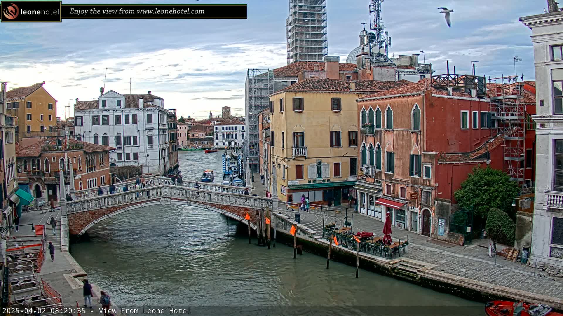 A stone bridge spans a canal in Venice, Italy, under a partly cloudy sky; several boats are visible on the water, and pedestrians are walking across the bridge and along the canal's edge.