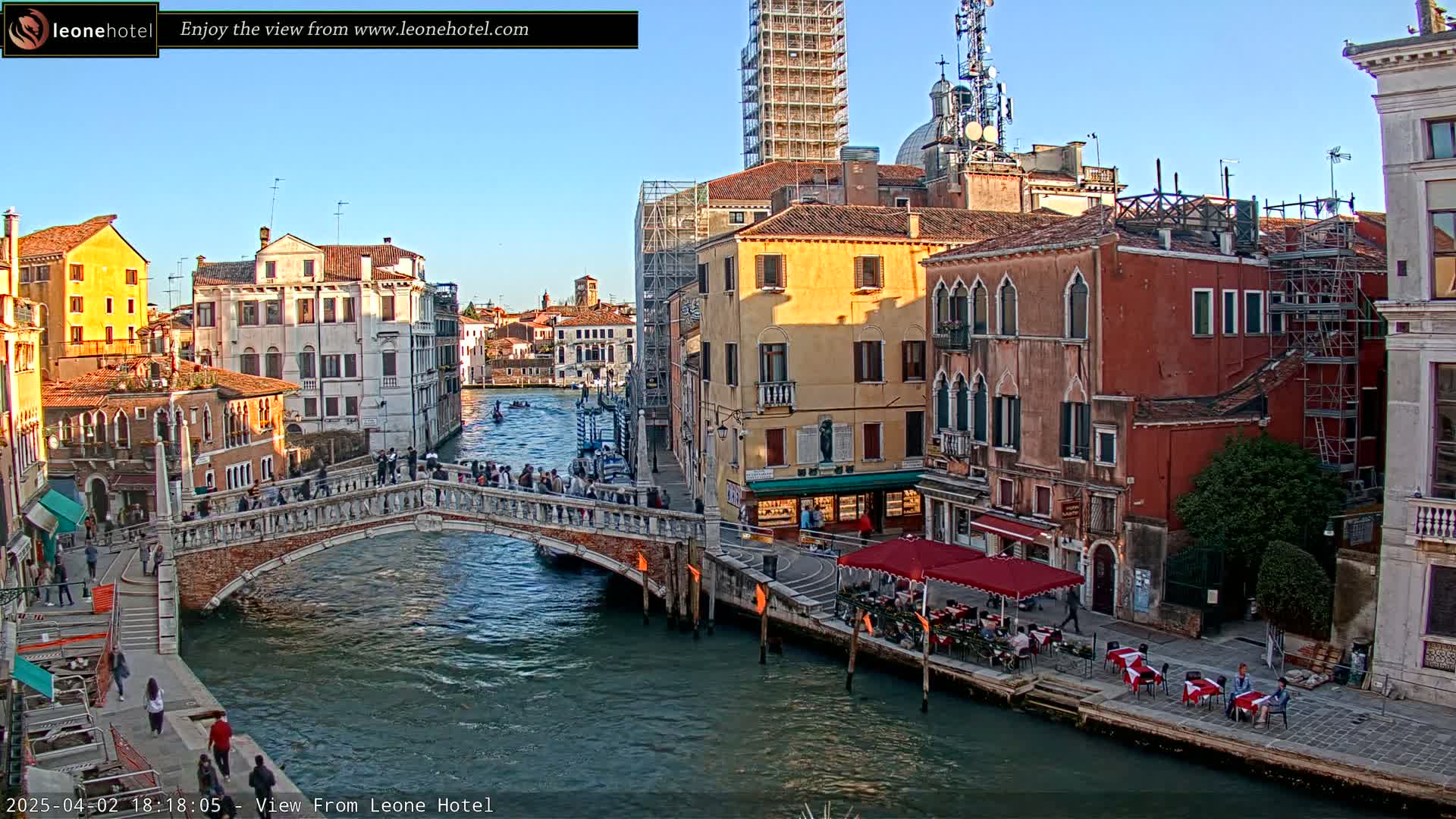 A Venetian canal scene under a clear, sunny sky shows a bridge with pedestrians crossing over it, colorful buildings lining the waterway, and outdoor seating at a canal-side restaurant.