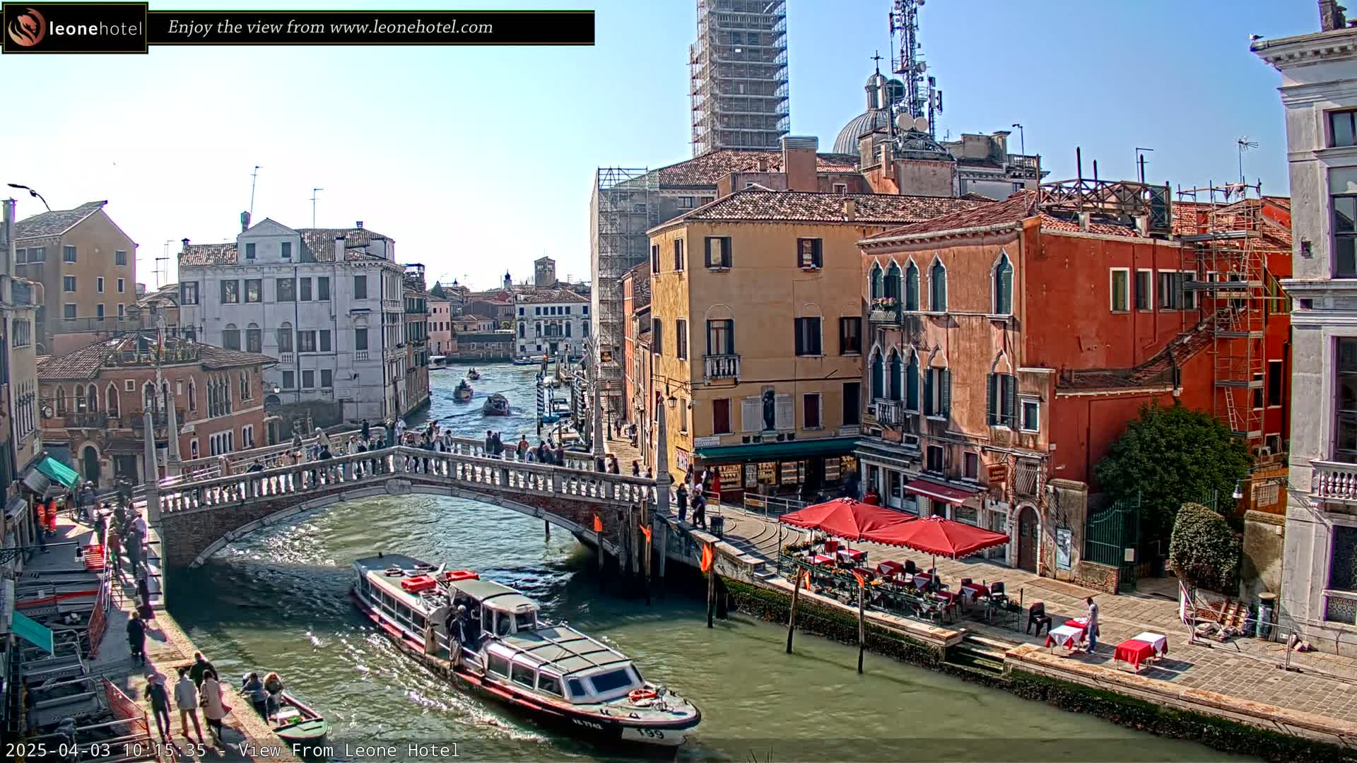 A sunny day in Venice shows a canal with a bridge, boats, and colorful buildings lining the waterway, one of which is under construction.