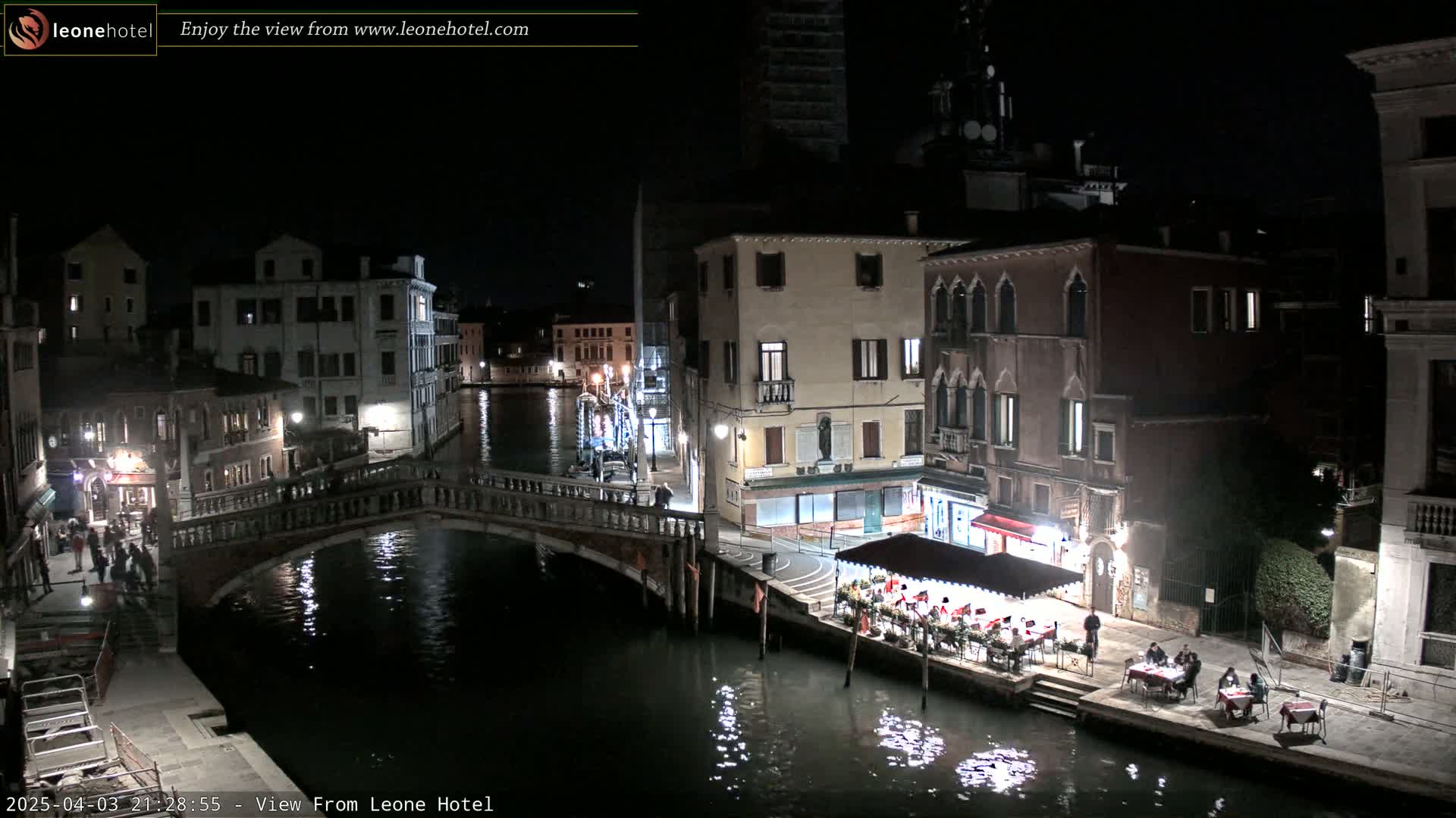 A nighttime view of a canal in Venice, Italy, showing a bridge, buildings, and several outdoor dining areas with people seated at tables.
