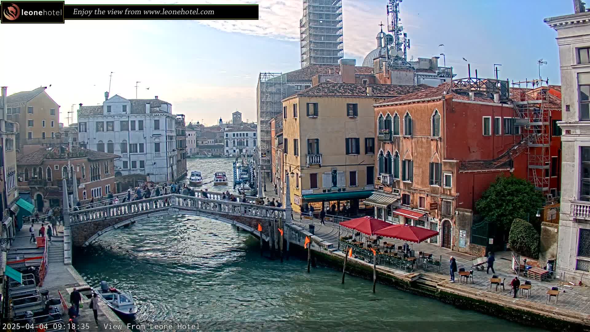 A stone bridge spans a canal in Venice, Italy, under a partly cloudy sky, with pedestrians crossing the bridge and colorful buildings lining the canal.