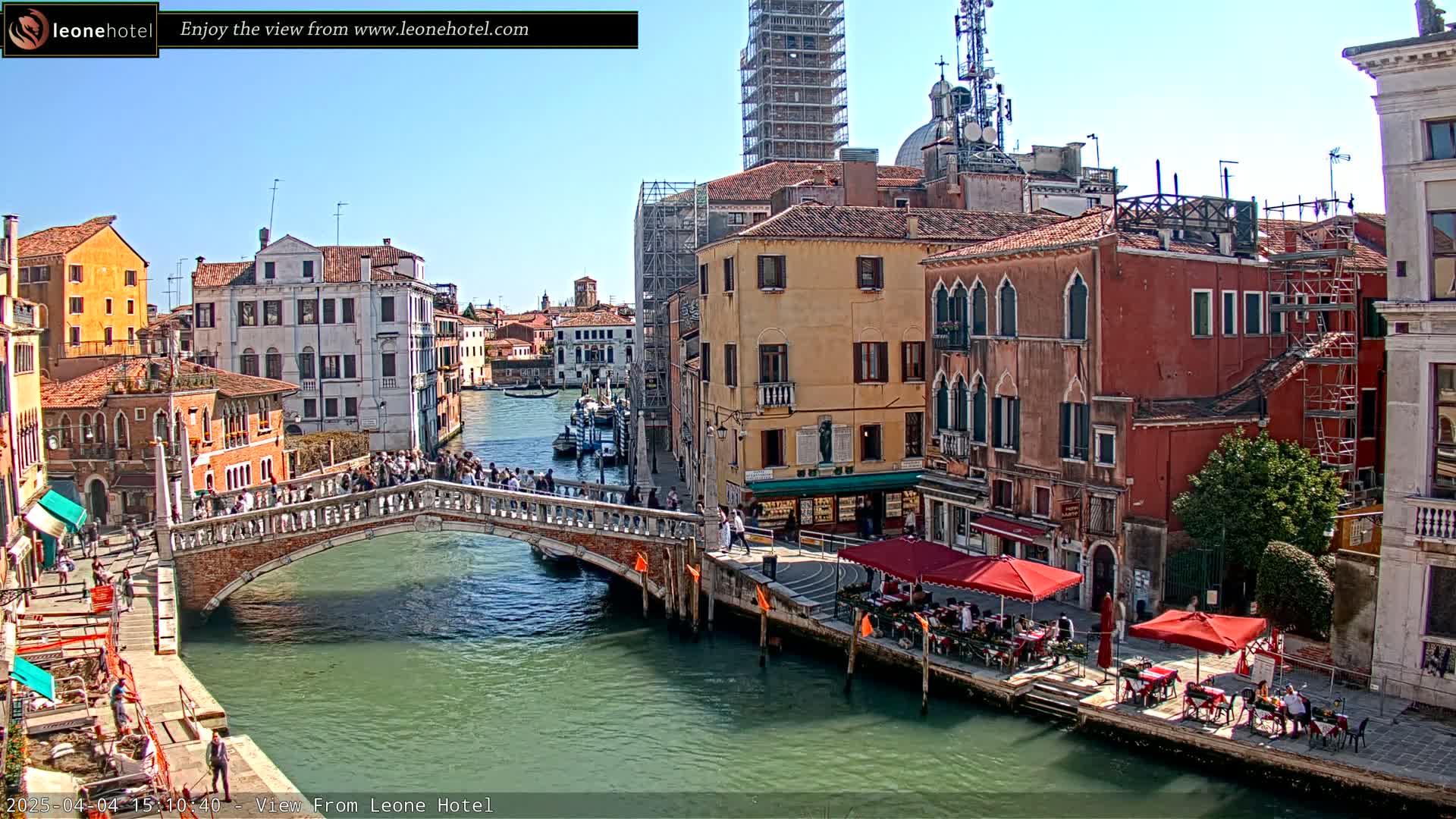 A sunny day in Venice shows a canal with a stone bridge crowded with pedestrians, and colorful buildings lining the waterway, some with outdoor cafes.