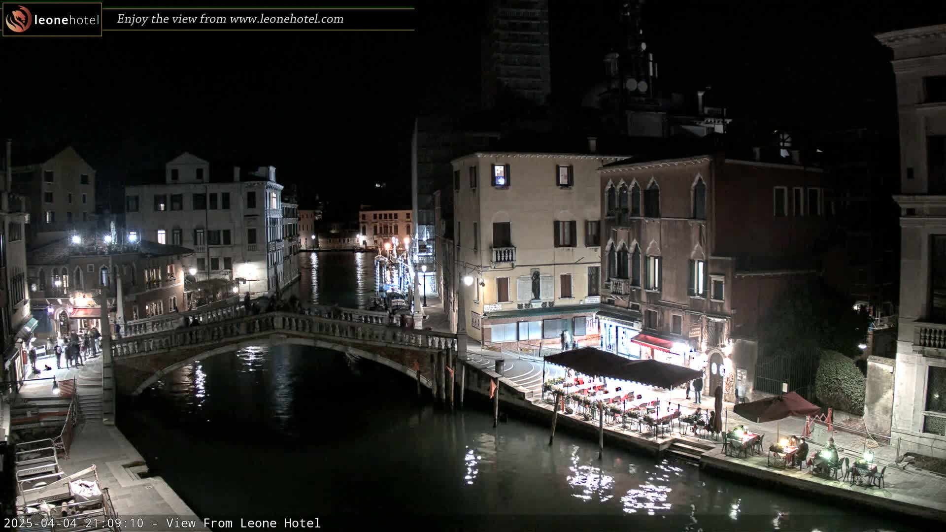 A nighttime view of a Venetian canal, showing bridges, buildings, and outdoor seating areas along the water's edge.