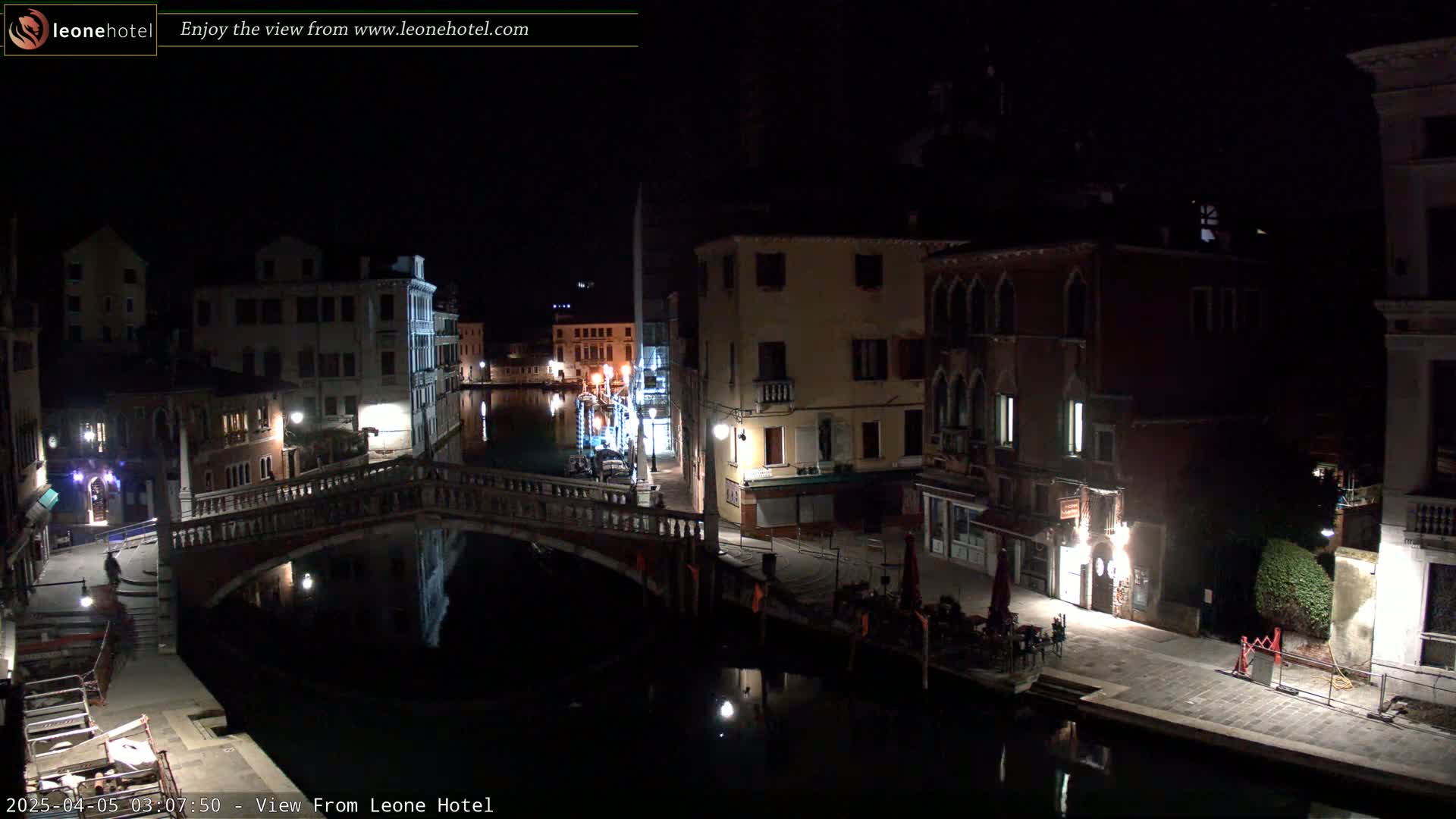 A nighttime view of a canal in Venice, Italy, shows several buildings lining the waterway, a bridge crossing the canal, and some outdoor seating areas.