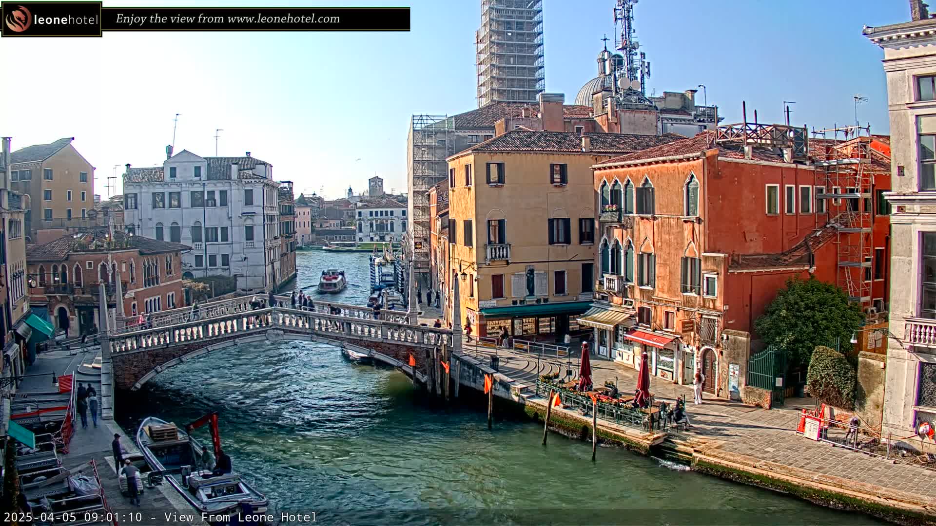 A sunny day in Venice shows a canal with a bridge, several buildings lining the waterway, and a few boats and people.