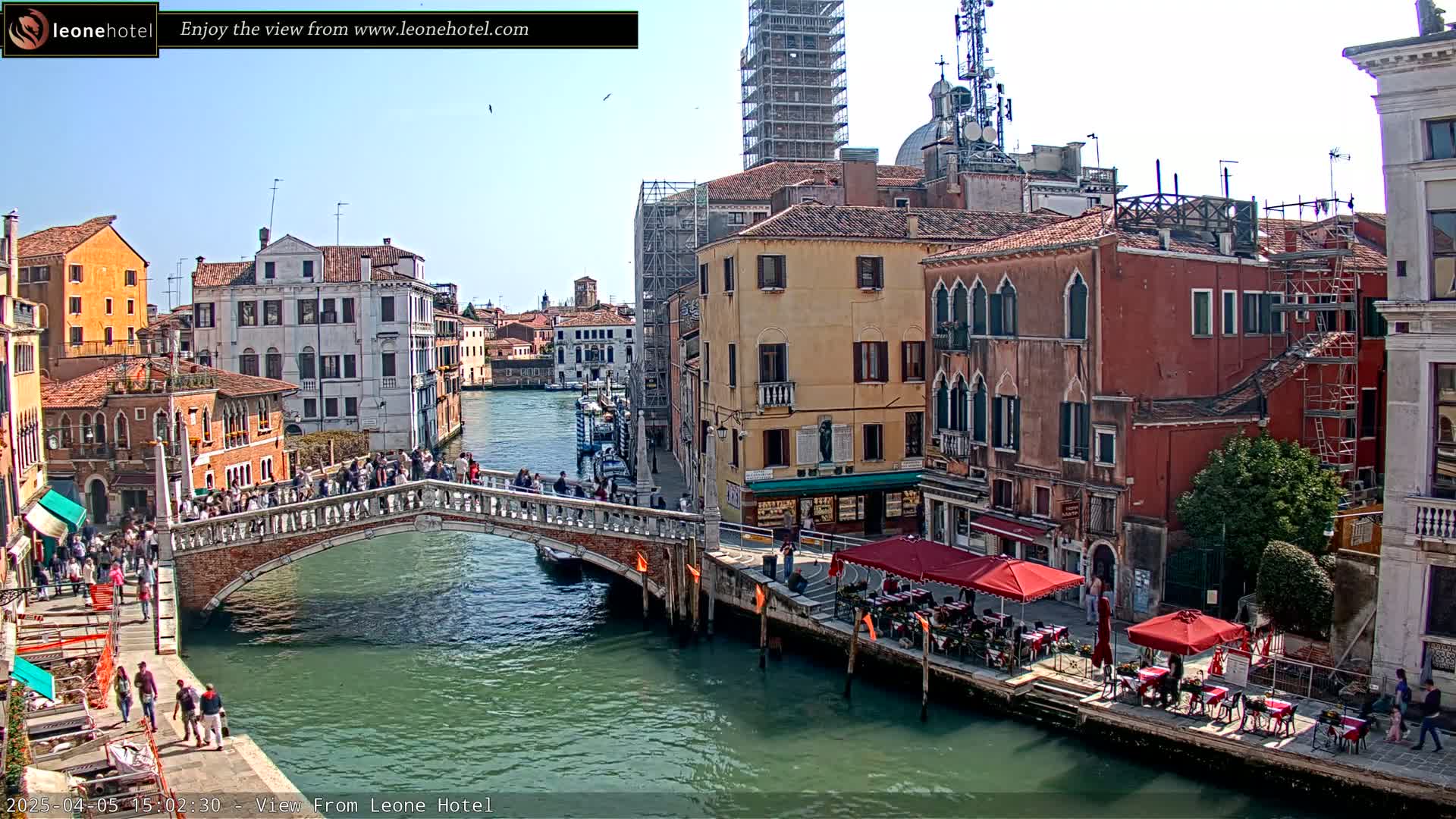 A sunny day in Venice shows a canal with a stone bridge crowded with people, lined with historic buildings, and featuring outdoor seating at a restaurant.