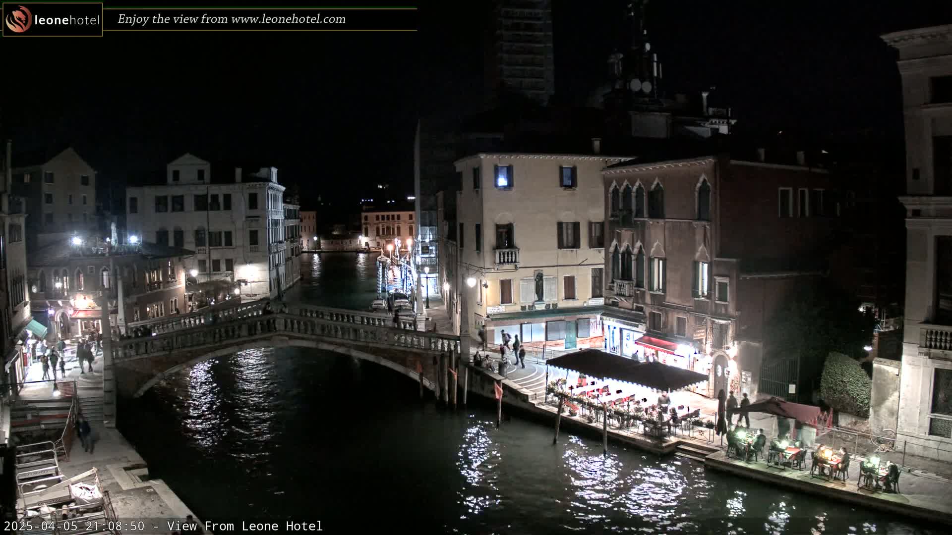 A nighttime view of a Venetian canal, showing bridges, buildings with lights, and people dining outdoors along the waterway under a clear, dark sky.