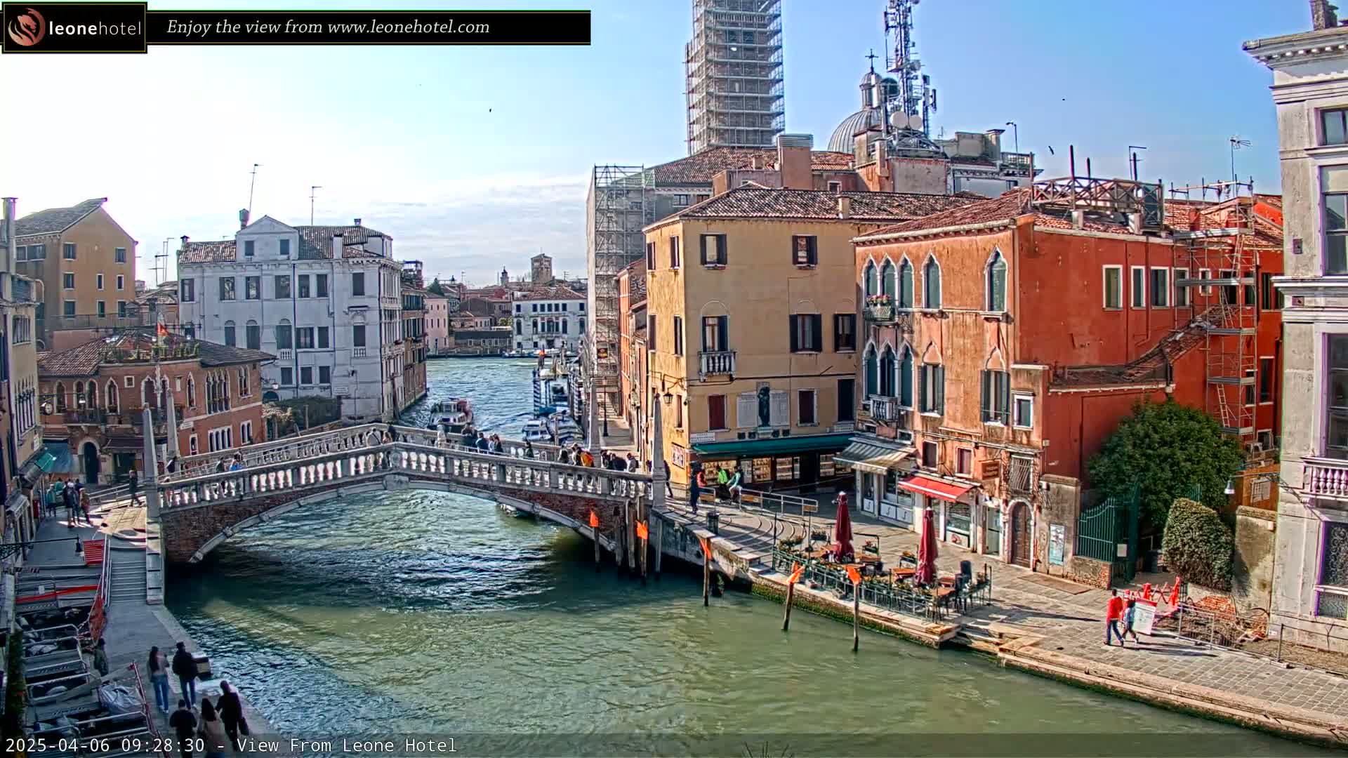 A sunny day in Venice shows a canal with a stone bridge, colorful buildings lining the waterway, and several boats and people.