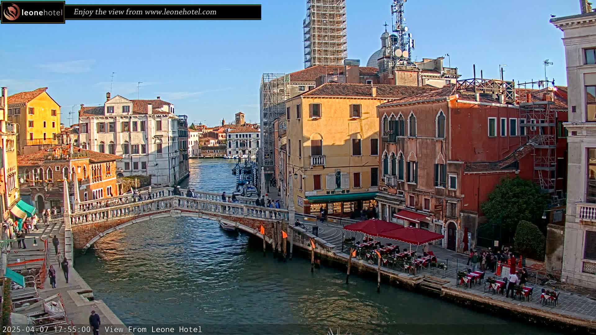A sunny day in Venice shows a canal with a stone bridge, colorful buildings lining the waterway, and outdoor seating at a restaurant.