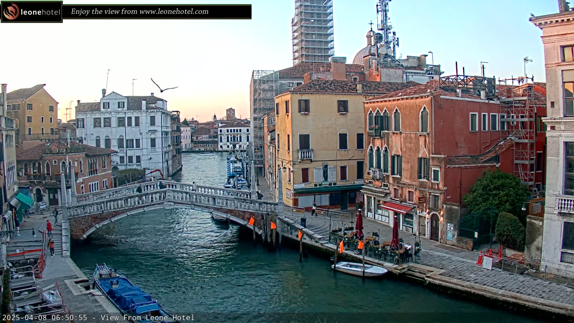 A tranquil canal scene in Venice, under a clear sky, shows colorful buildings, a stone bridge, and several boats, with some outdoor seating at a cafe.