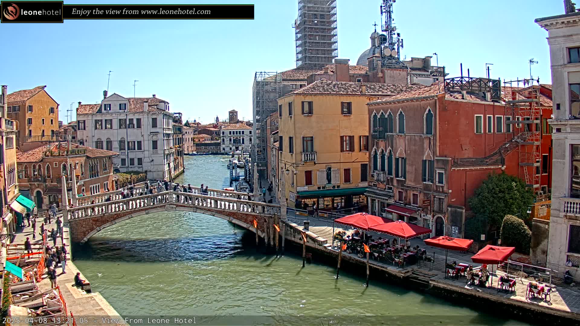 A sunny day in Venice shows a canal with a stone bridge, colorful buildings lining the waterway, and several outdoor cafes with red umbrellas.