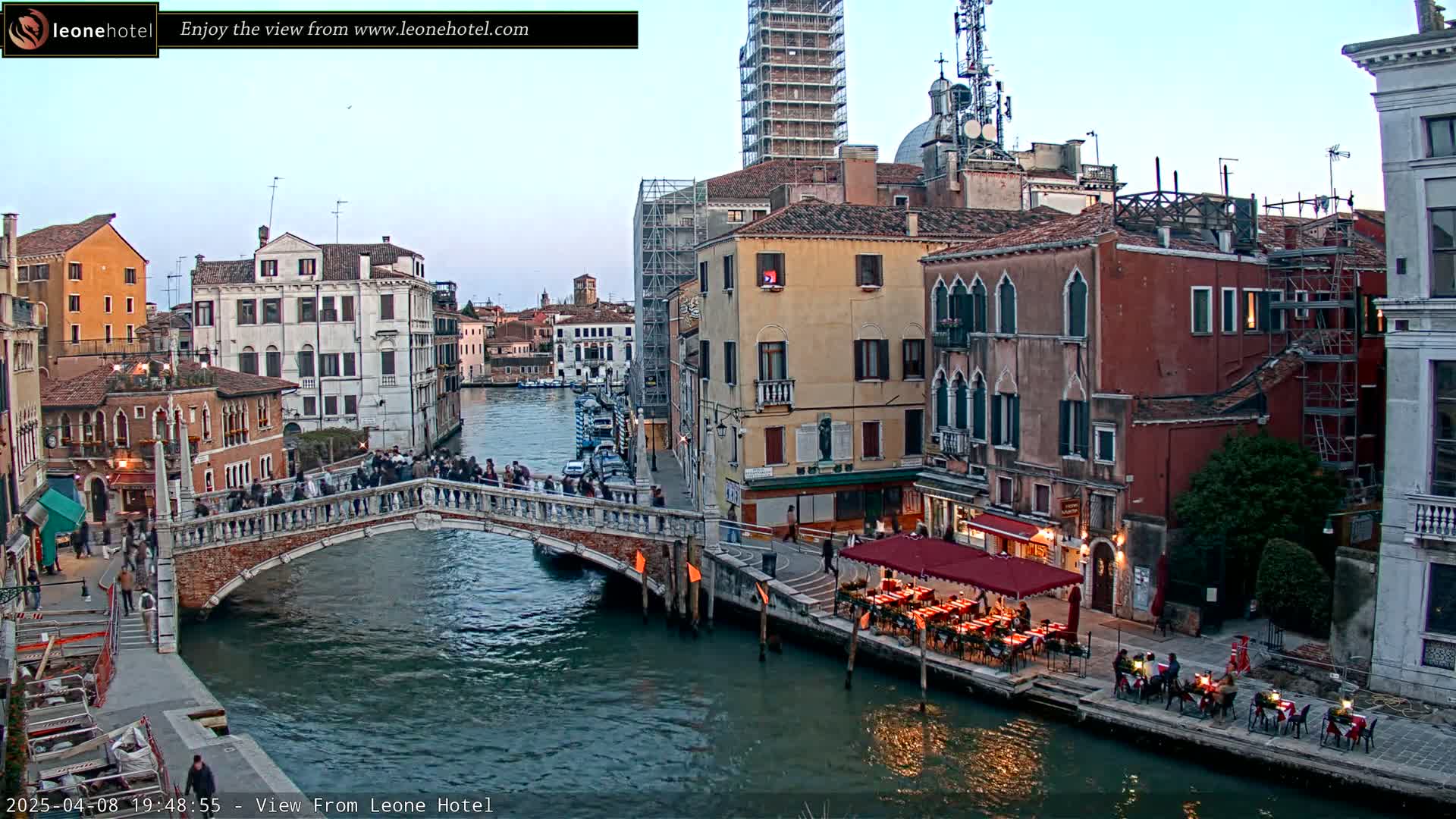 A bridge over a canal in Venice, Italy, at dusk, with people walking across it and several buildings lining the waterway, one of which has outdoor seating.