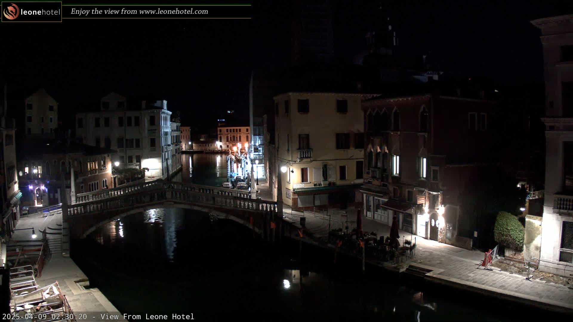 A nighttime view of a canal in Venice, Italy, shows a bridge with buildings and some outdoor seating along the waterway under a clear, dark sky.
