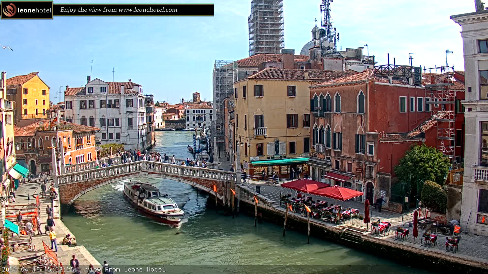 A sunny day in Venice shows a canal with a boat passing under a stone bridge, lined with buildings and outdoor seating.