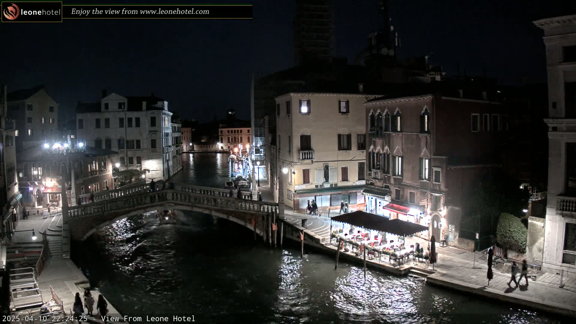A nighttime view of a canal in Venice, Italy, showing a bridge, buildings, and a restaurant with outdoor seating under a canopy.