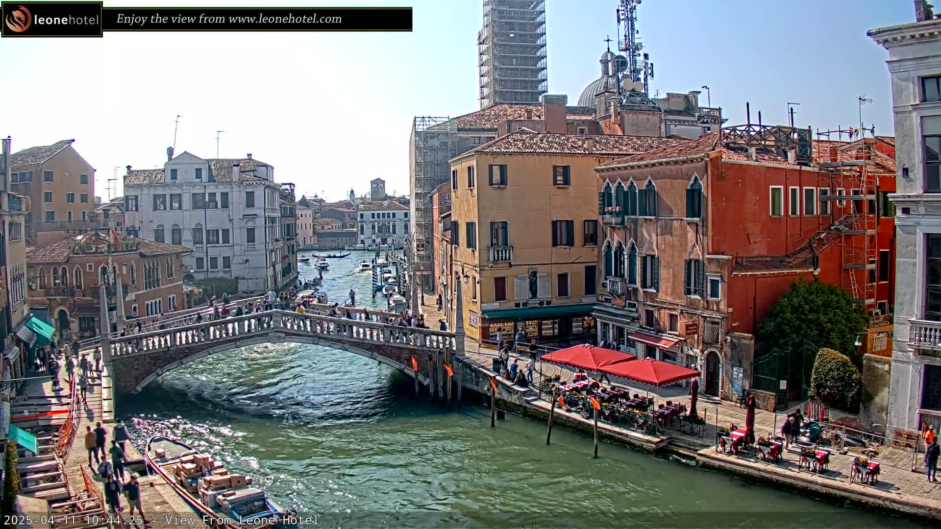 A sunny day in Venice shows a canal with boats, a stone bridge crowded with people, and colorful buildings lining the waterfront, some undergoing construction.