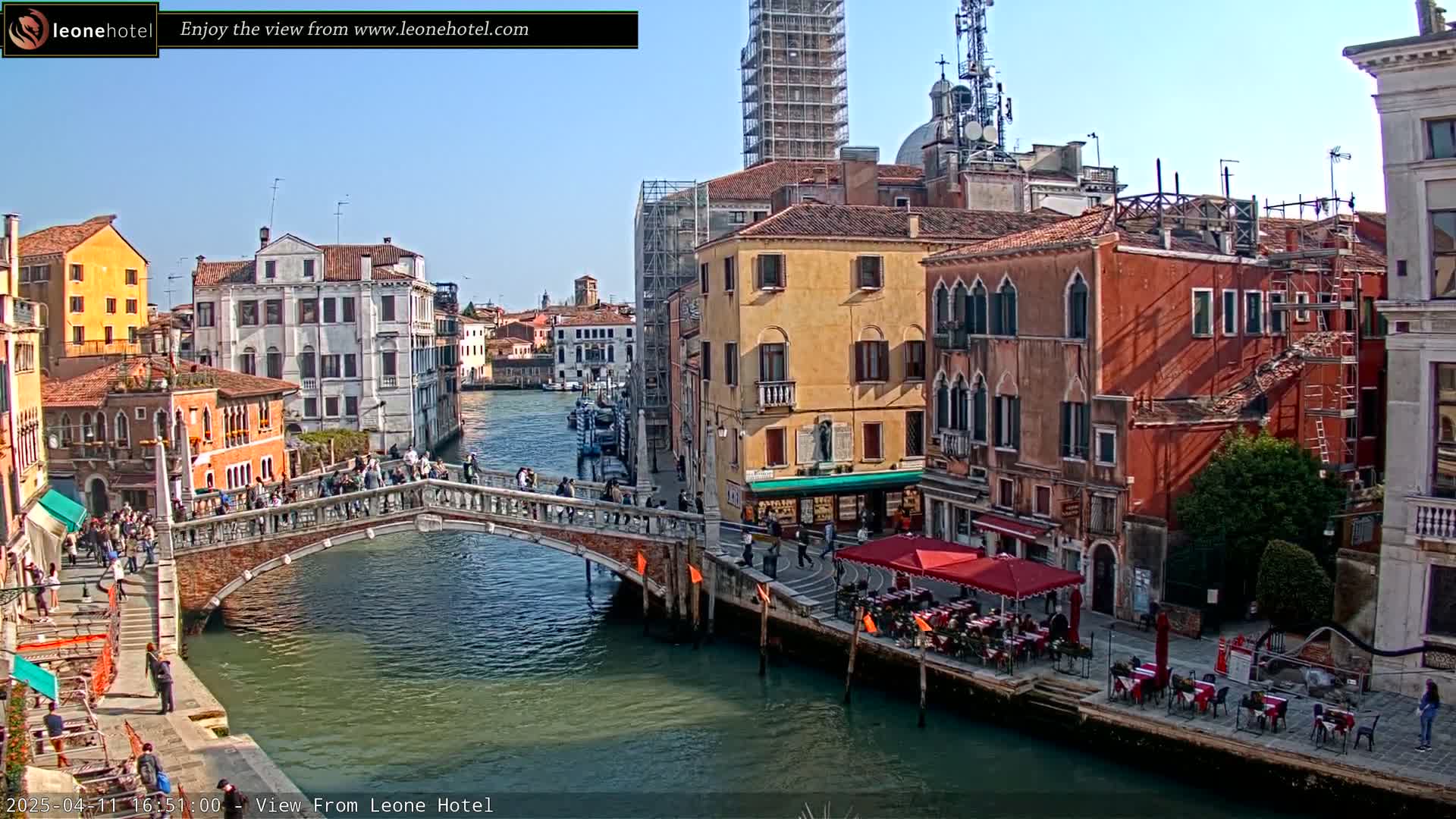 A sunny day in Venice shows a canal with a bridge crowded with people, colorful buildings lining the waterway, and outdoor seating at cafes.