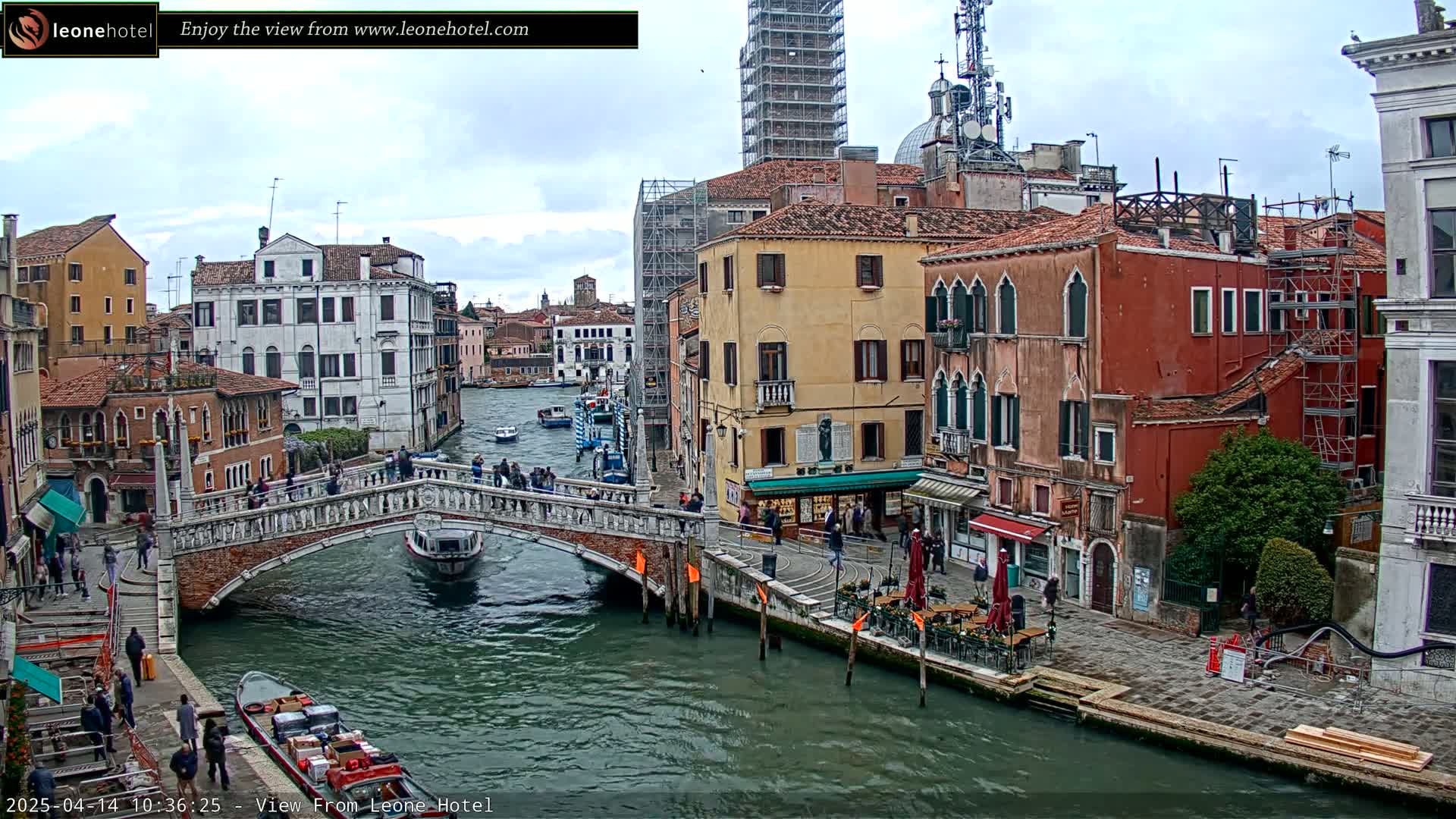 A canal in Venice, under an overcast sky, is viewed from a bridge, showing several boats and pedestrians along the waterway and buildings lining the canals.