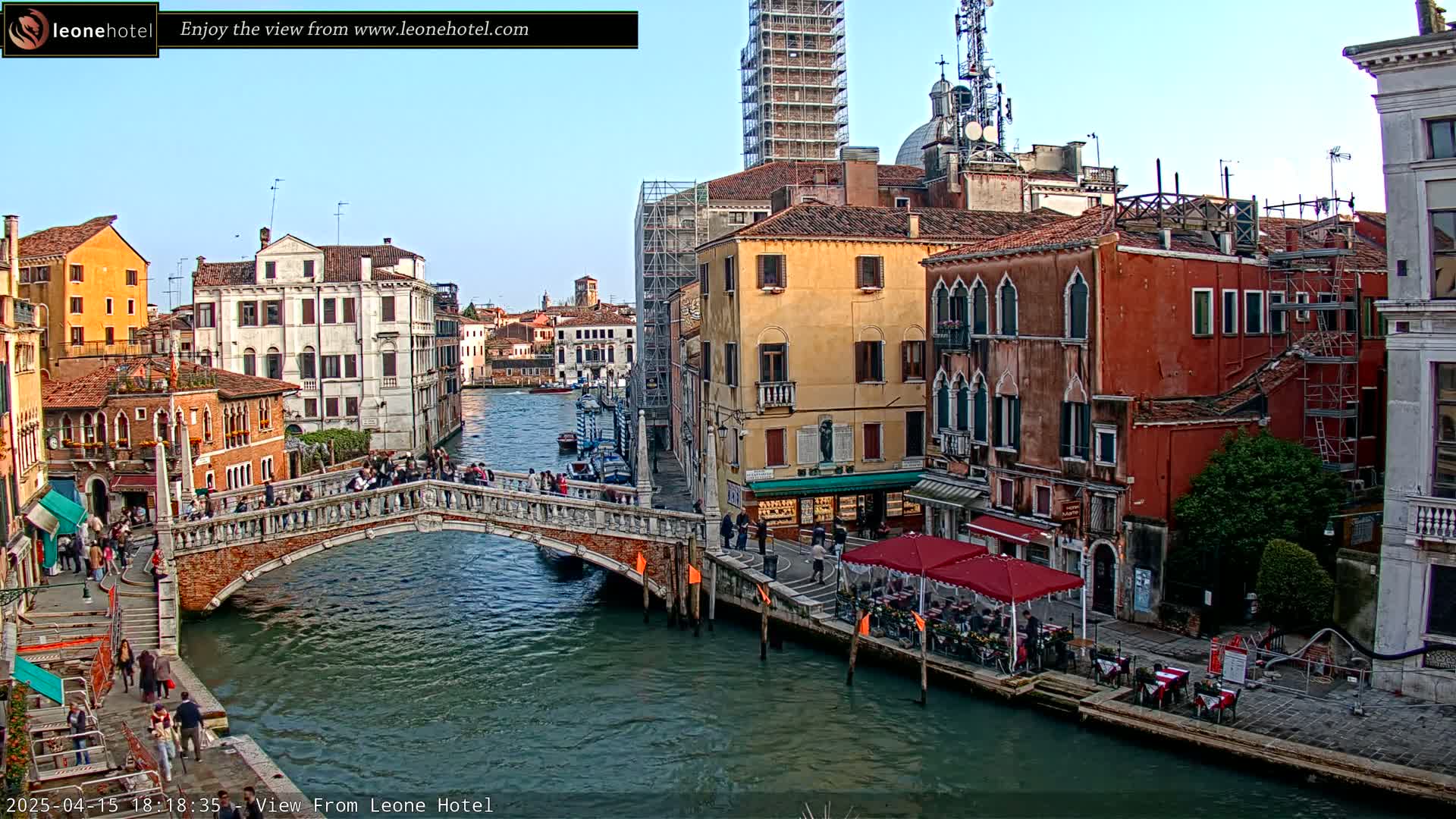 A stone bridge crosses a canal in Venice, Italy, under a clear sky, with colorful buildings lining the waterway and people walking on the bridge and sidewalks.