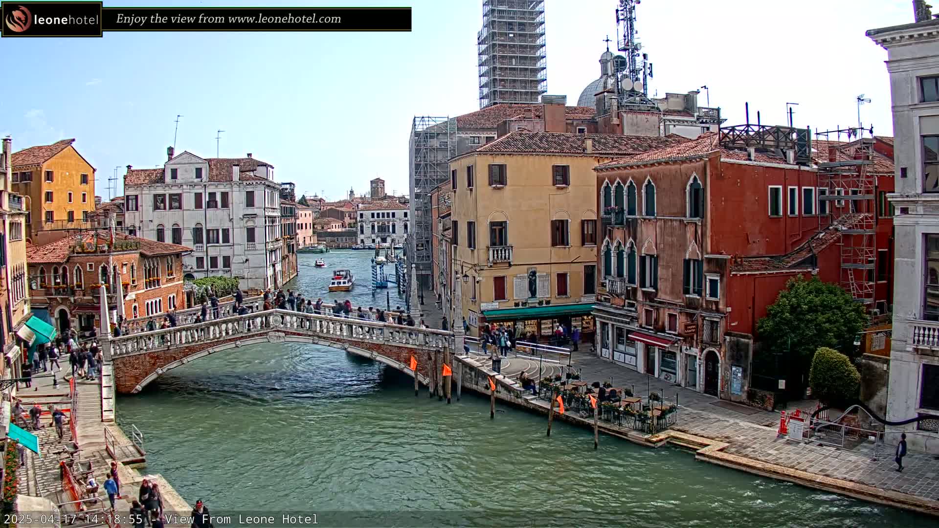 A stone bridge spans a canal in Venice, Italy, under a mostly clear sky, with colorful buildings lining the waterway and people walking on the bridge and the canal-side walkways.