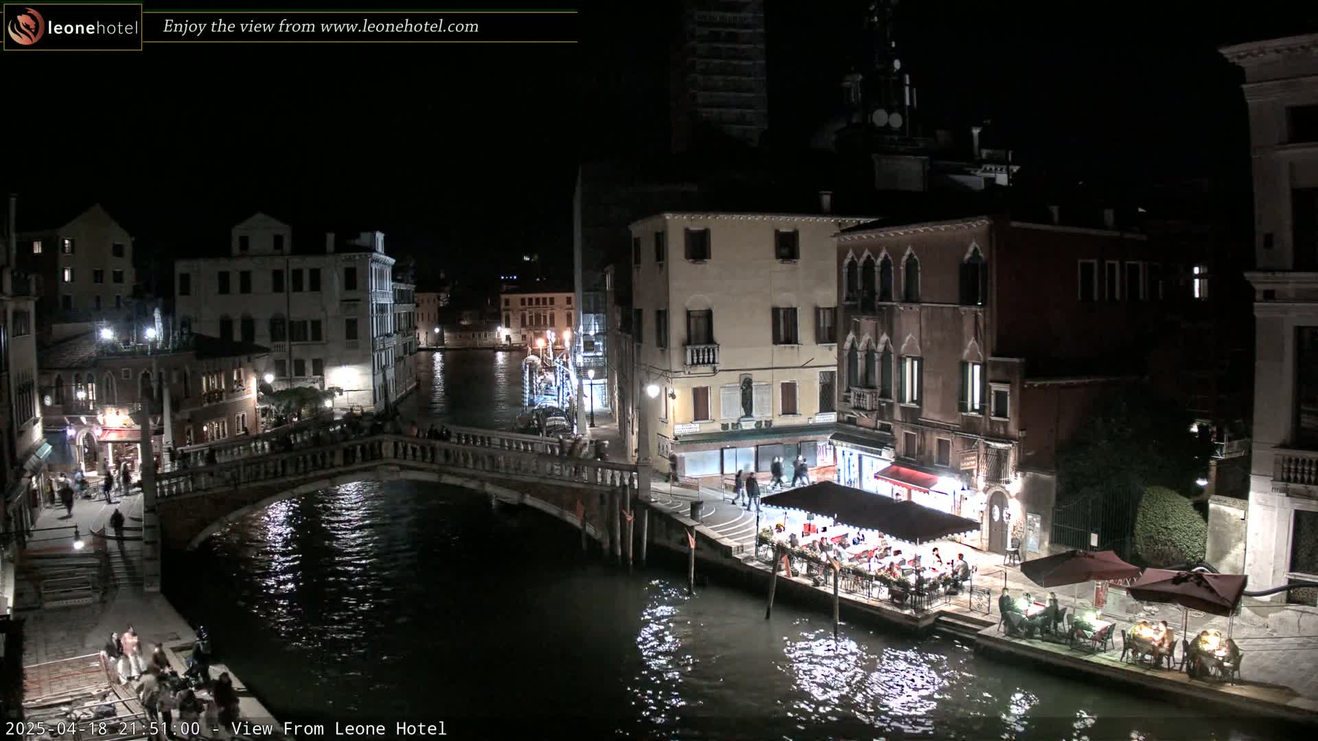 A nighttime view of a Venetian canal, showing bridges, buildings, and people dining outdoors at waterside restaurants.