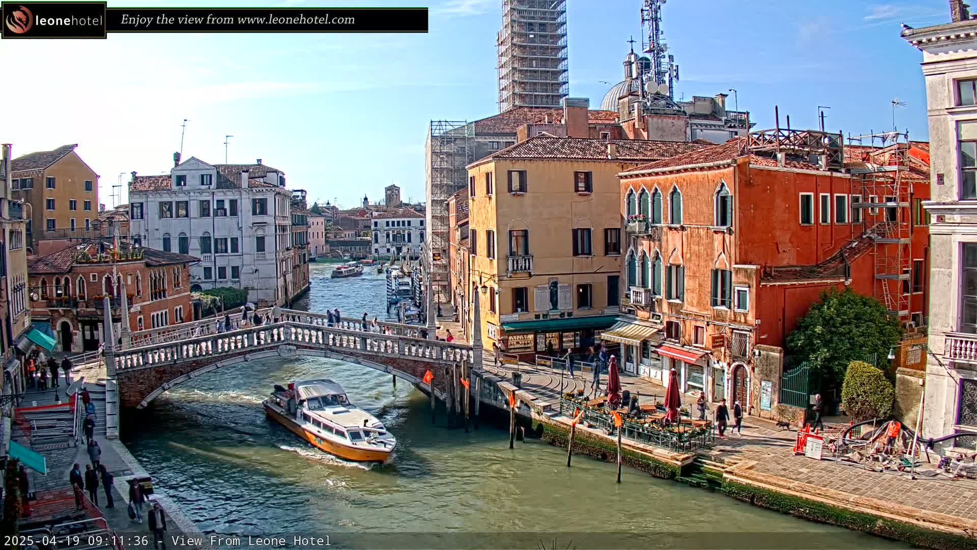 A sunny day in Venice shows a canal with a bridge, boats, and colorful buildings lining the waterway.