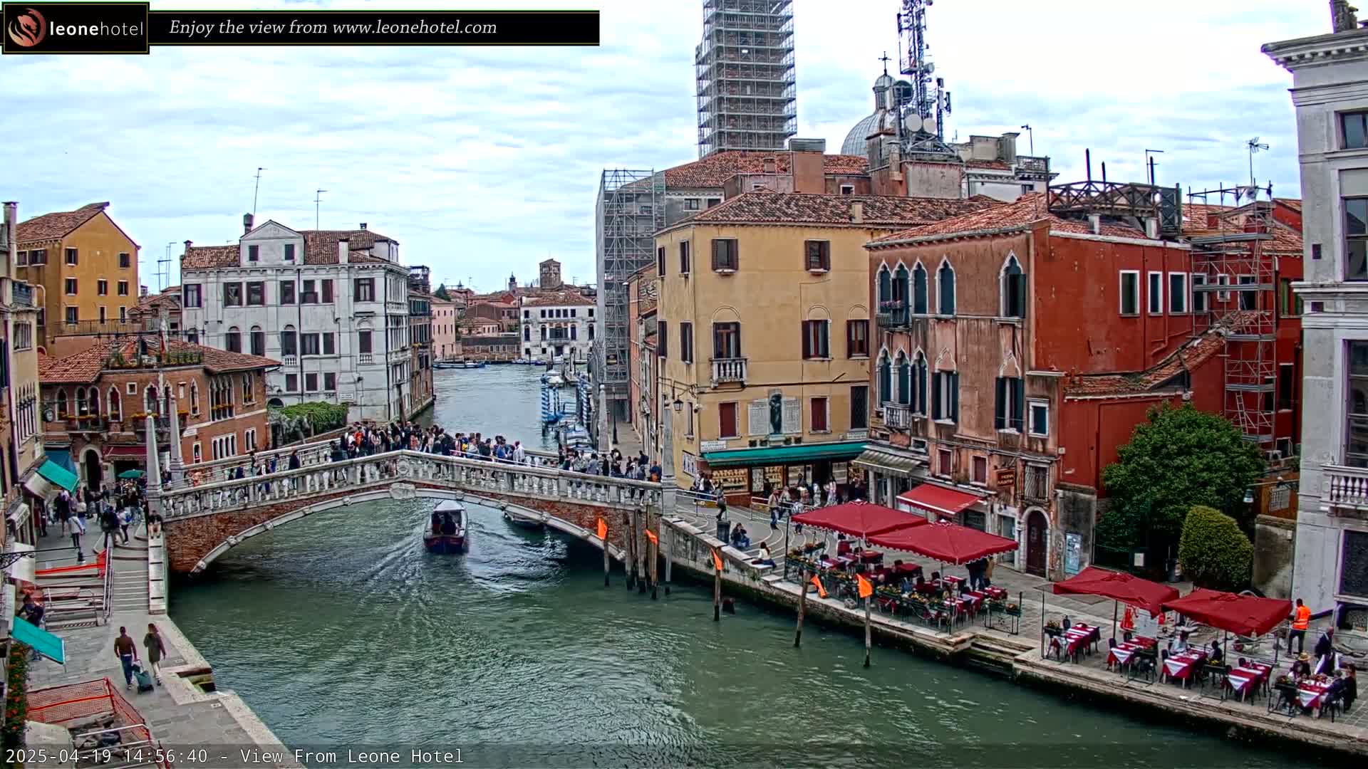 A bridge crowded with people spans a canal in Venice, Italy, under an overcast sky; several buildings line the canal, some with outdoor seating.