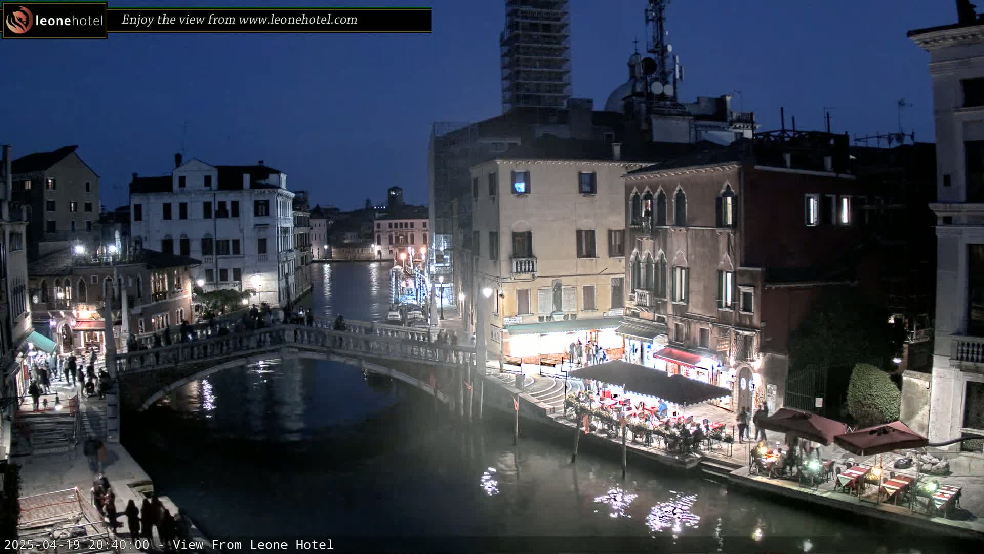 A nighttime view of a Venetian canal, showing bridges, buildings with lights, and people along the waterway under a dark sky.