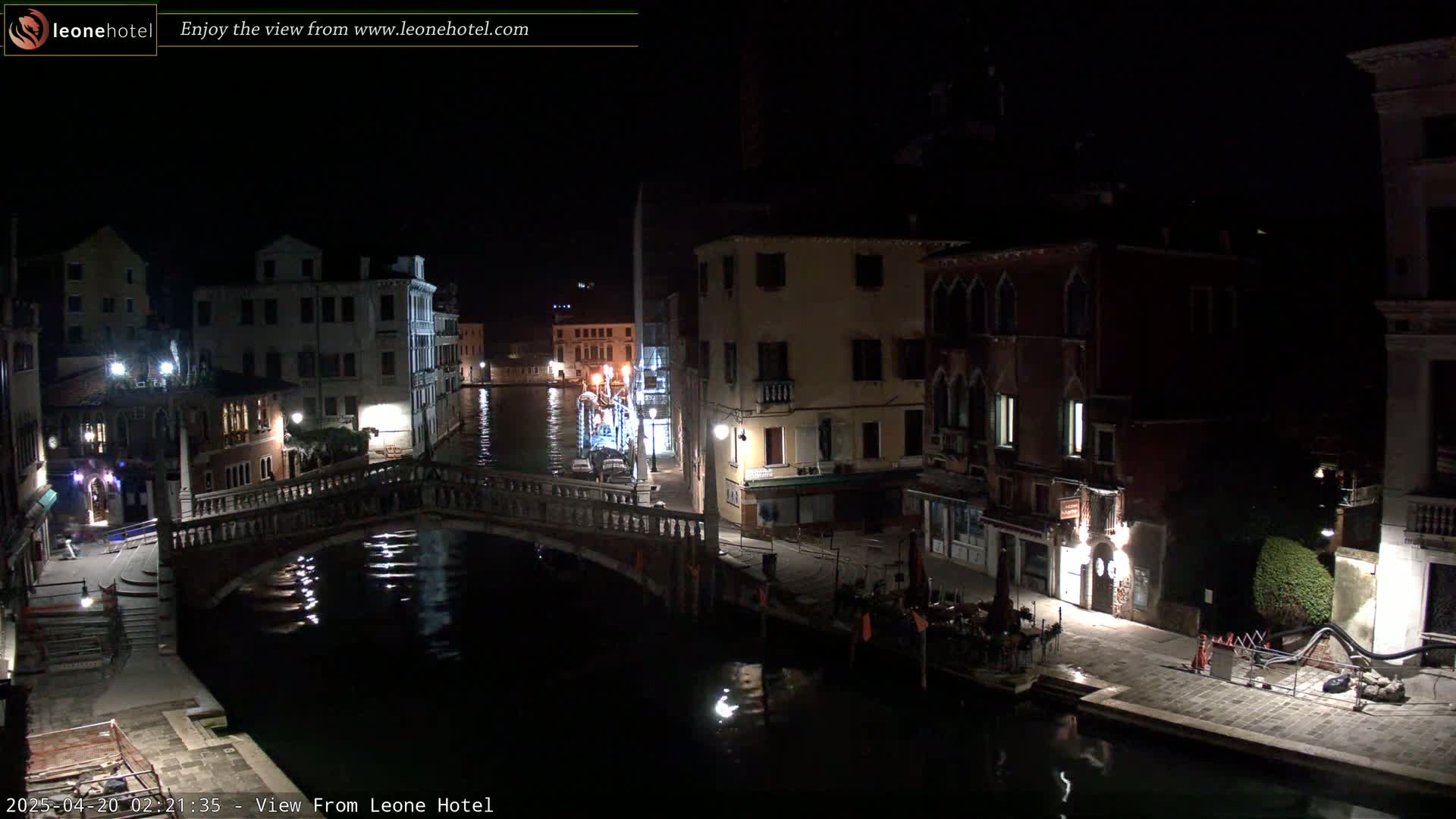 A nighttime view of a canal in Venice, Italy, shows a bridge and illuminated buildings lining the waterway.