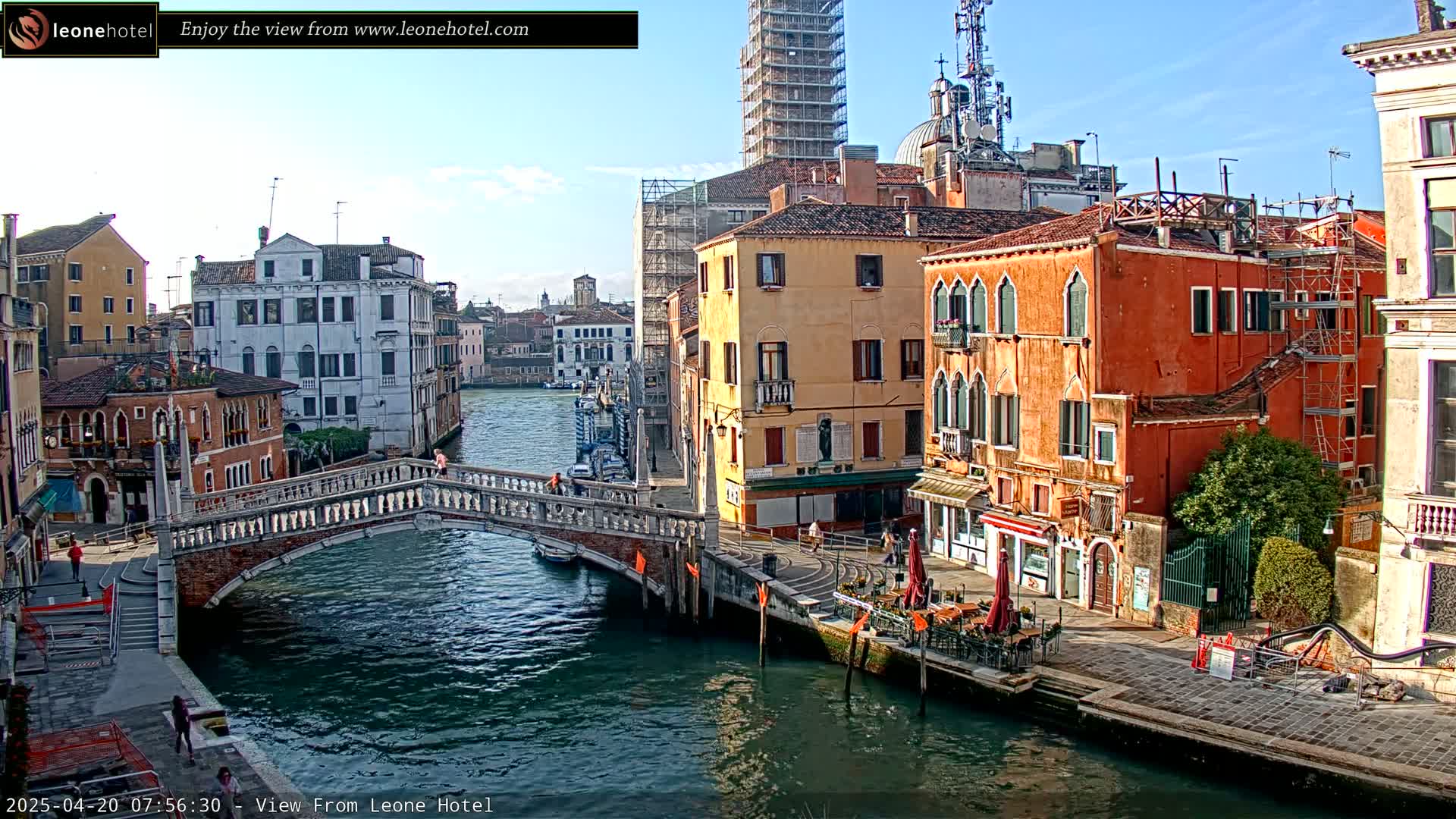 A sunny day in Venice shows a canal with colorful buildings lining its banks, a stone bridge spanning the water, and a few pedestrians.