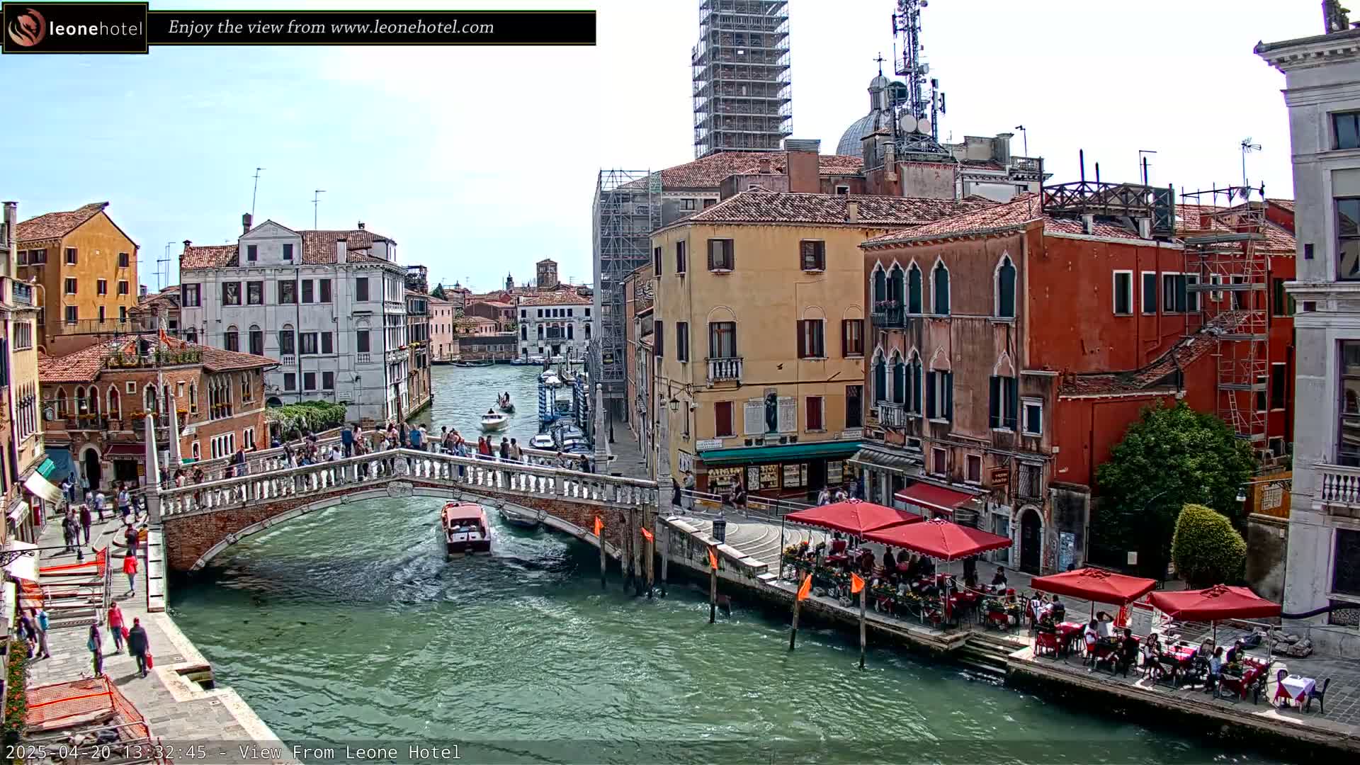 A sunny day in Venice shows a canal with a bridge full of people, boats on the water, and outdoor cafes lining the waterway next to colorful buildings, one of which is under construction.