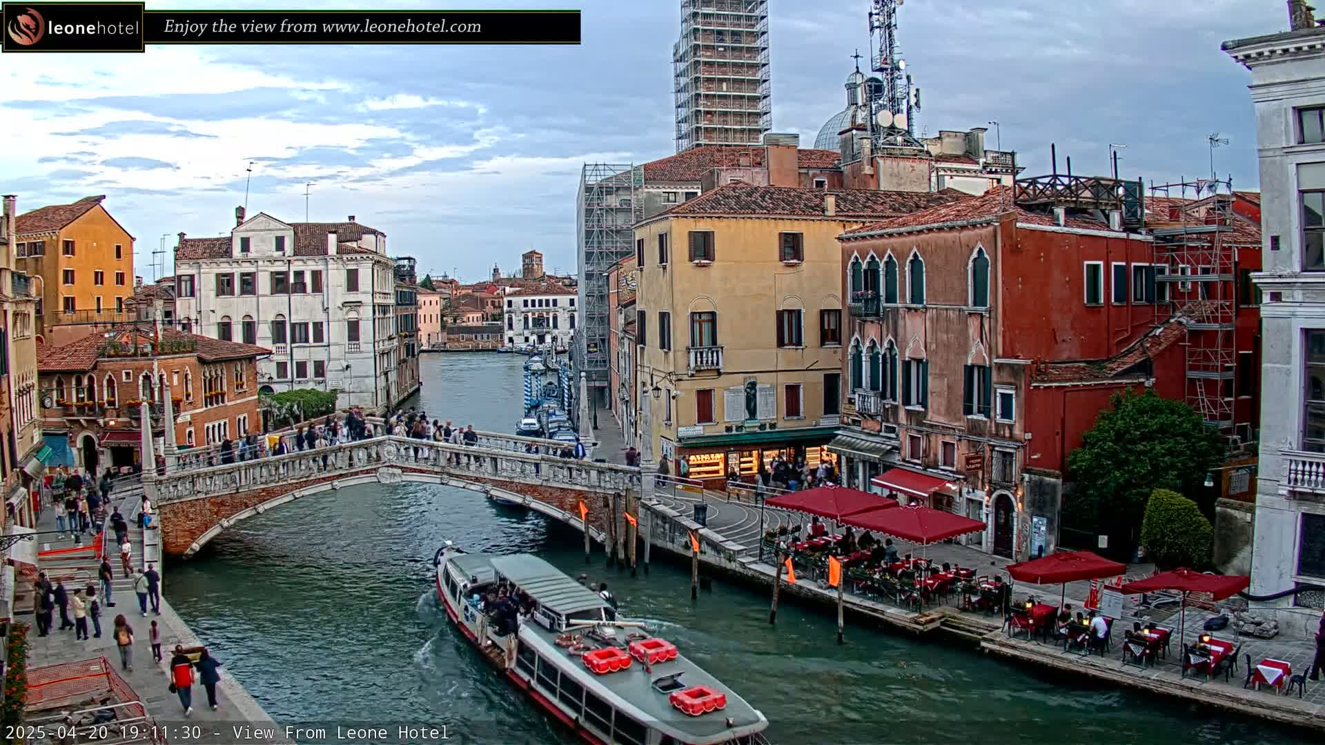 A Venetian canal scene under a partly cloudy sky shows a bridge with pedestrians, colorful buildings lining the waterway, and a boat carrying passengers.