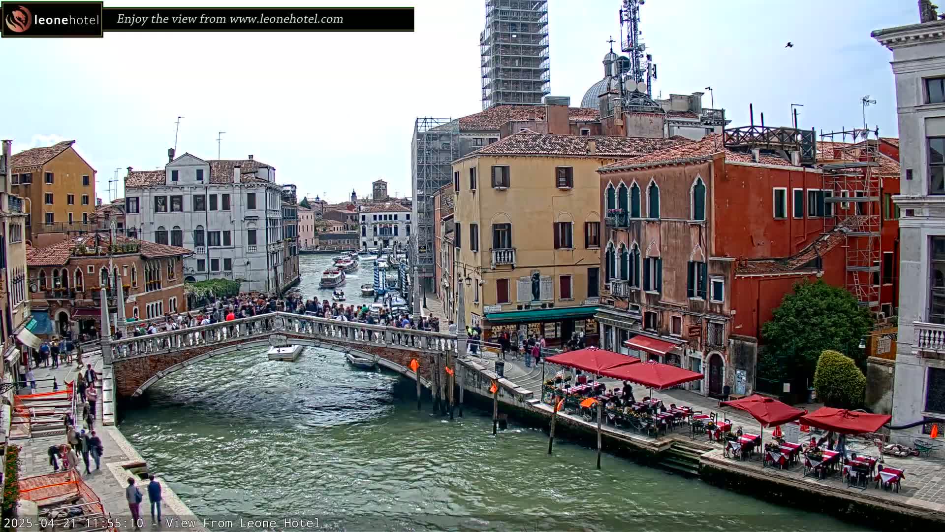 A canal in Venice, Italy, with a bridge crowded with people, boats on the water, and buildings lining the canal under a partly cloudy sky.