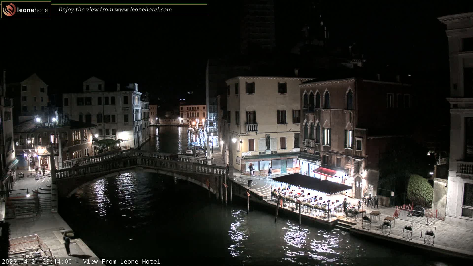 A nighttime view of a Venetian canal, showing bridges, buildings, and an outdoor restaurant with patrons, under a clear, dark sky.